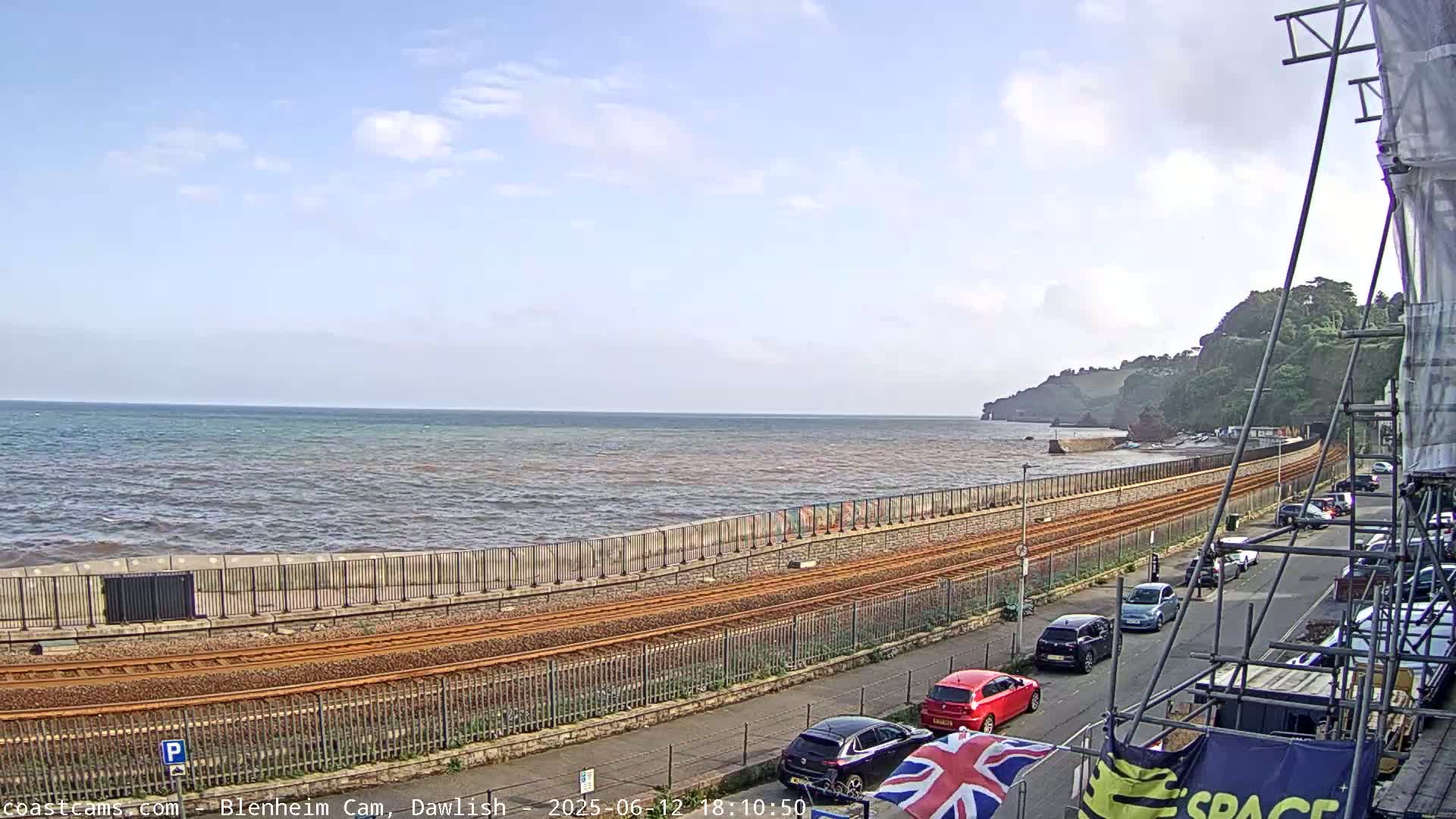 A slightly choppy sea meets a coastal railway line beside a road with parked cars, under a partly cloudy sky.