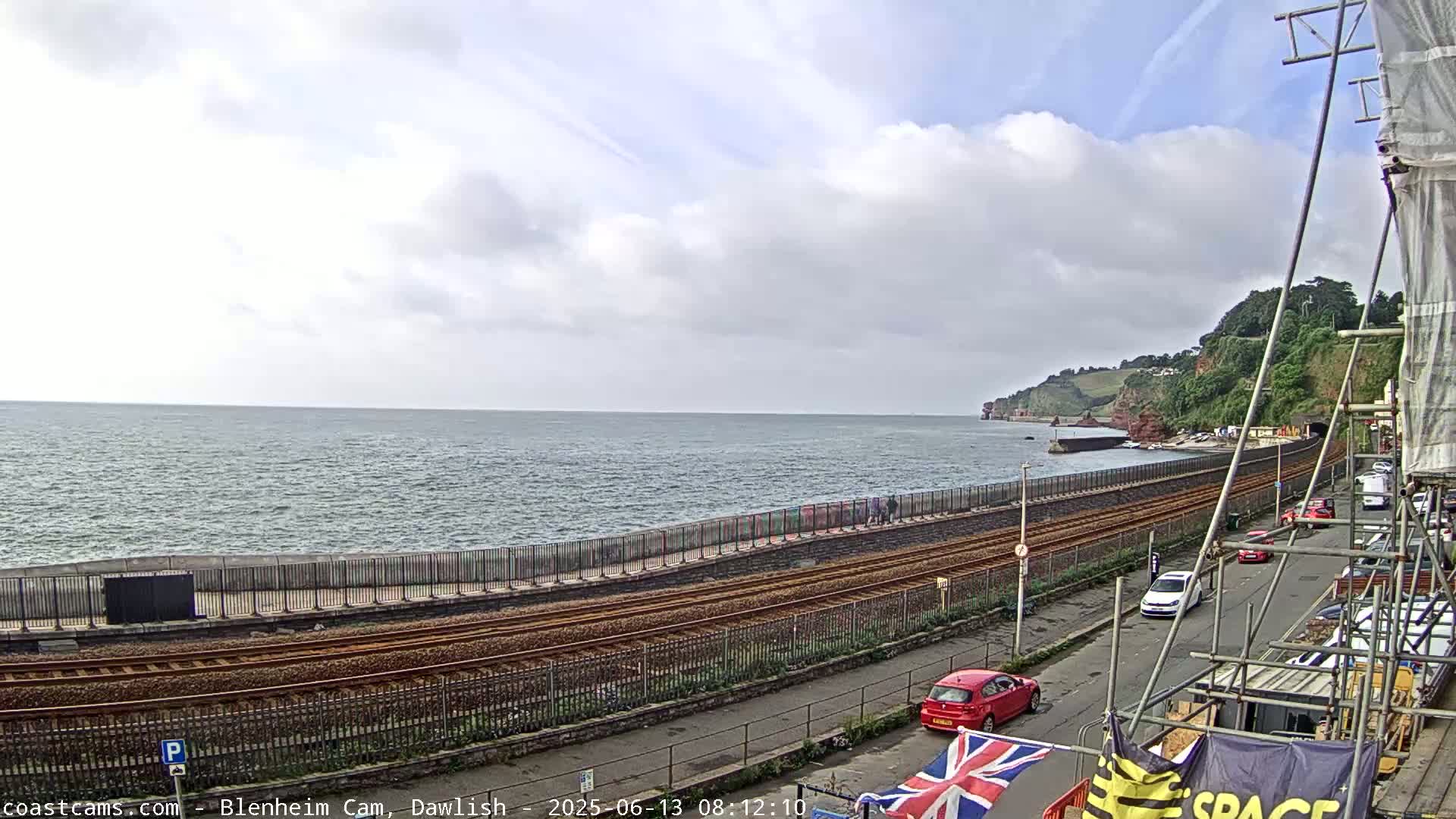 A coastal scene shows a partly cloudy sky over a calm sea, with railroad tracks running along a seawall next to a road with parked cars, and a green hillside in the distance.