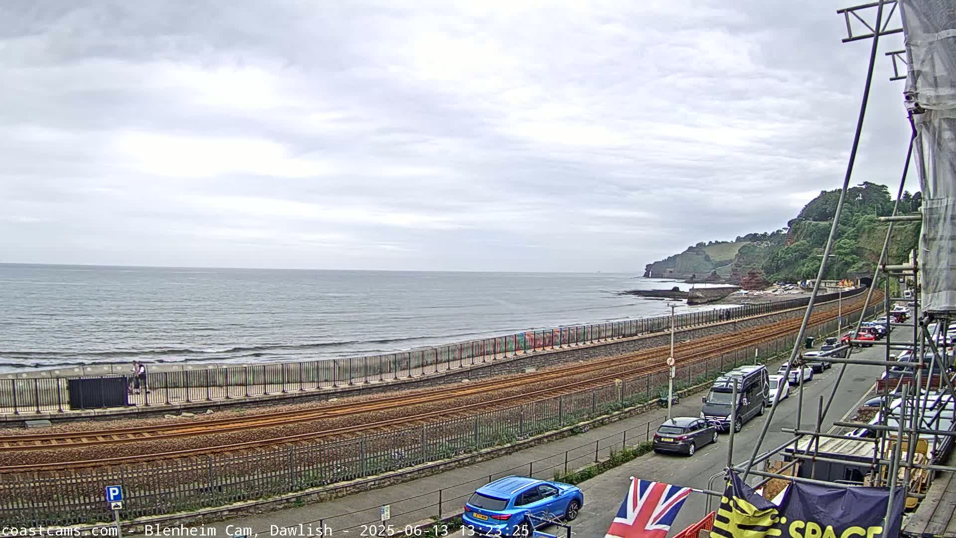 A mostly cloudy day reveals a coastline view of a beach with a railway track running alongside it, cars parked on a street, and a partially obscured building under construction.