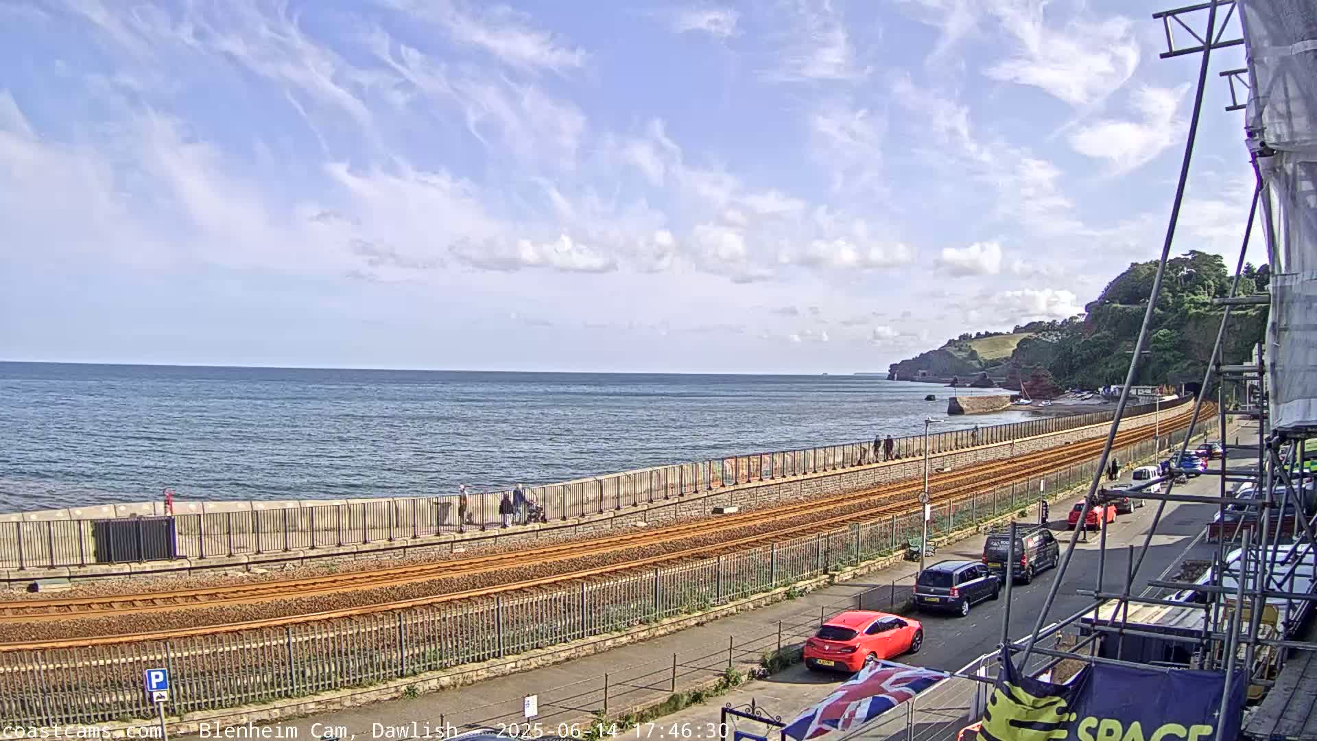 A seaside scene under a partly cloudy sky shows a train track running alongside a seawall with people walking along it, cars parked on a street next to it, and a body of water with a distant shoreline.