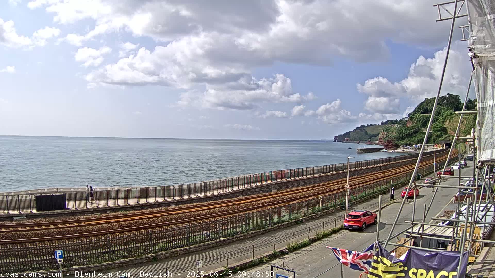 A partly cloudy day shows a coastal scene with train tracks running alongside a seawall next to the ocean, with a few cars parked nearby and a building under construction.