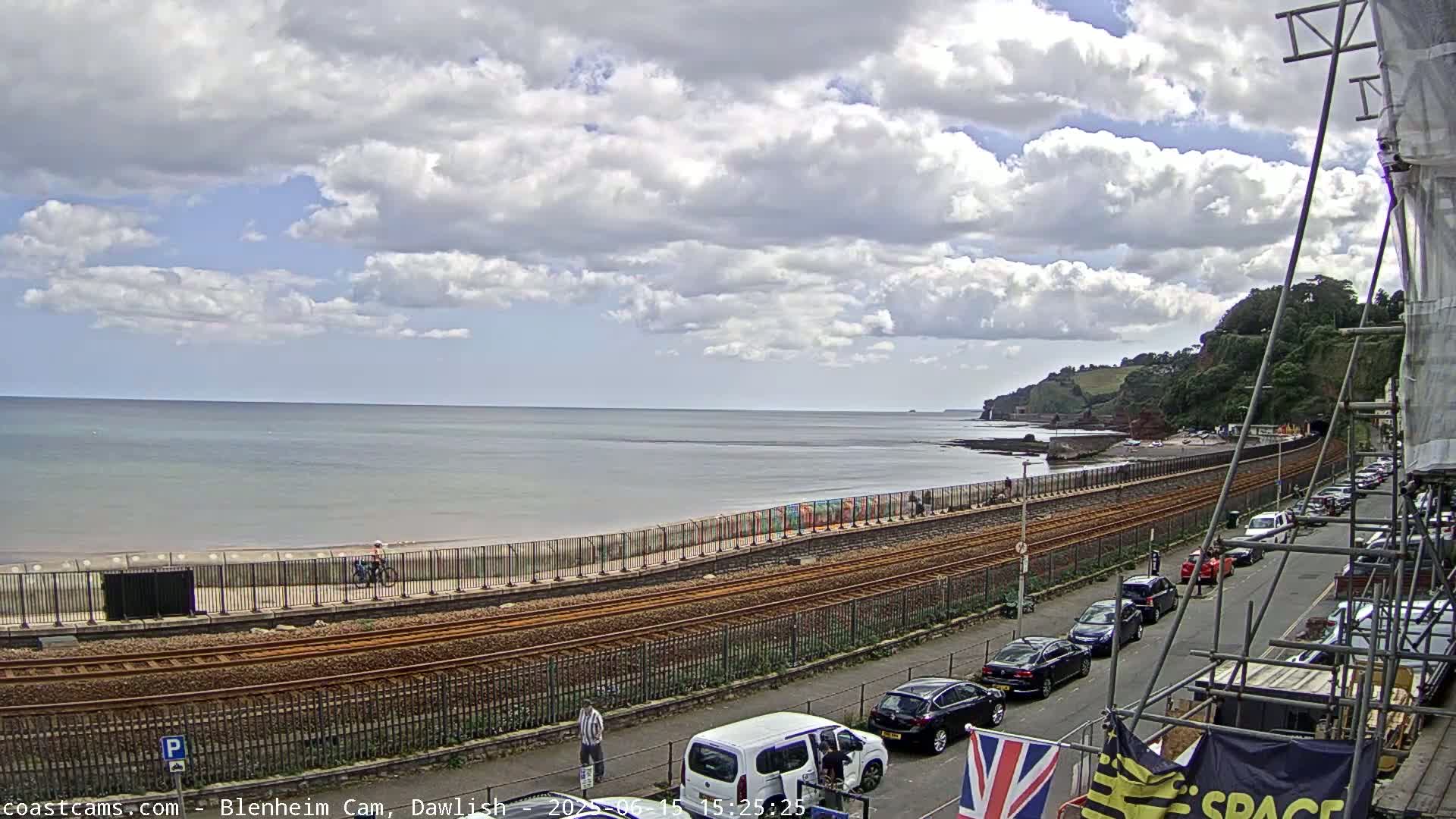 A partly cloudy day shows a beach, ocean, railway tracks, parked cars along a road, and a seaside cliff in the distance.