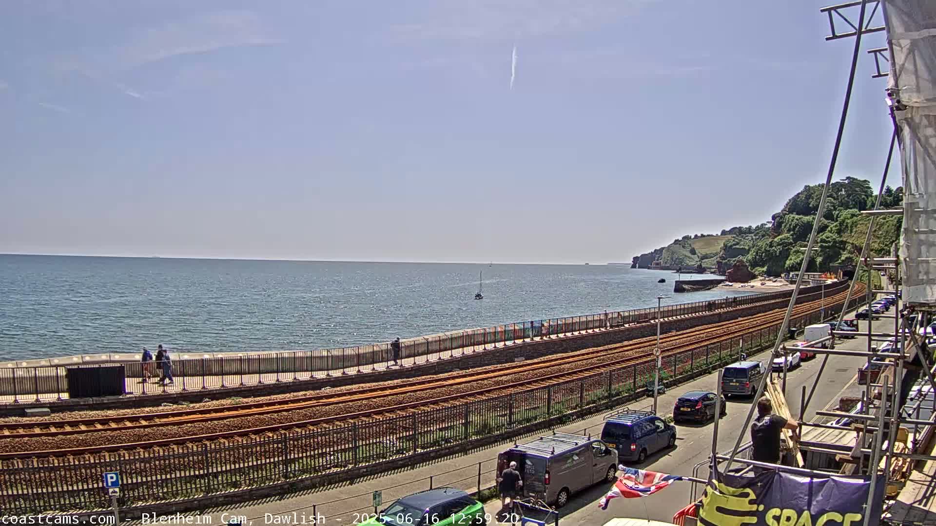 A seaside scene on a sunny day shows a calm ocean, a rail line running alongside a paved walkway, and parked cars along a road next to the tracks.
