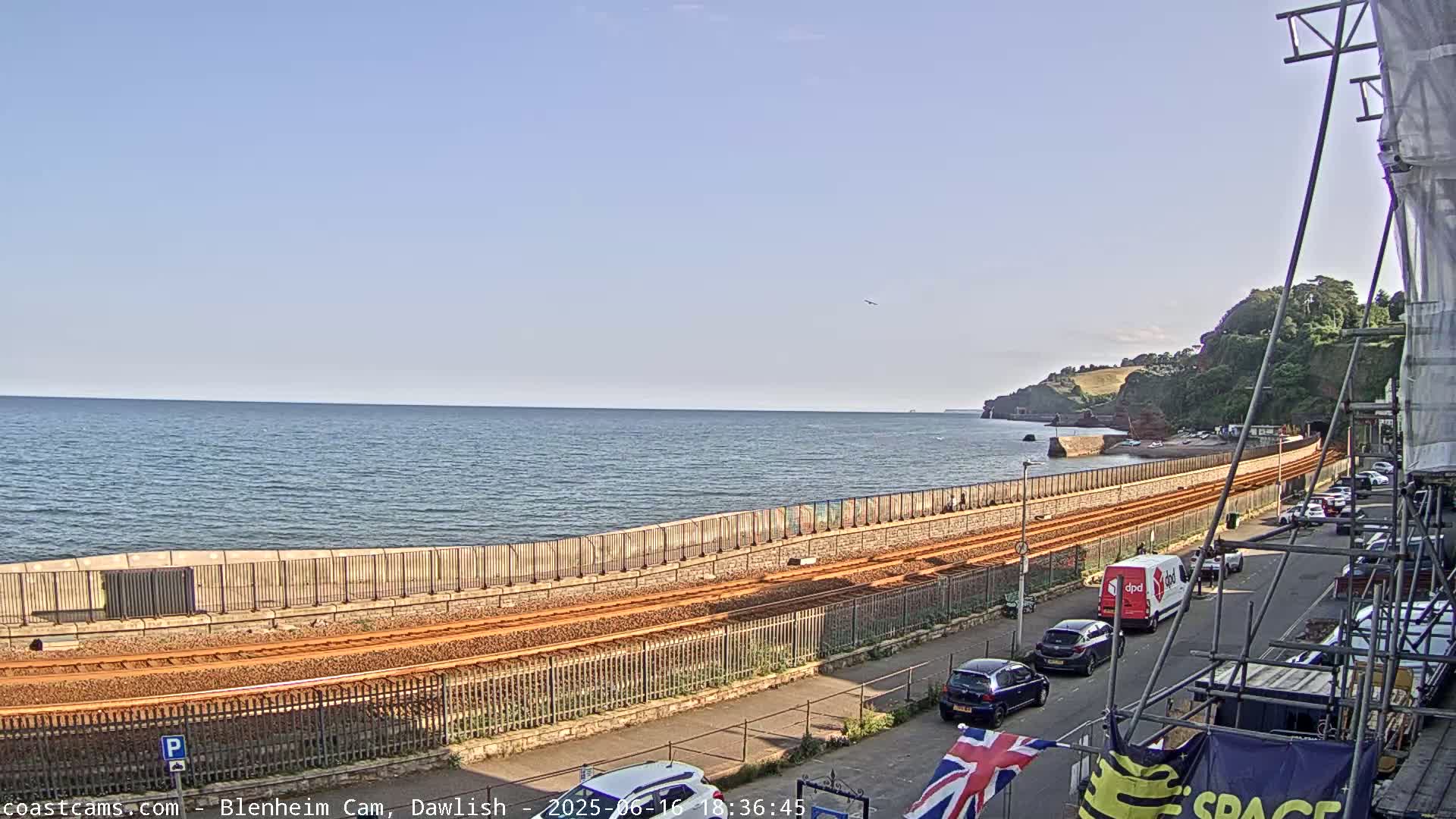 A coastal scene on a sunny day shows a train track running alongside a sea wall, with cars parked along a street next to it, and a calm ocean stretching to a distant headland.