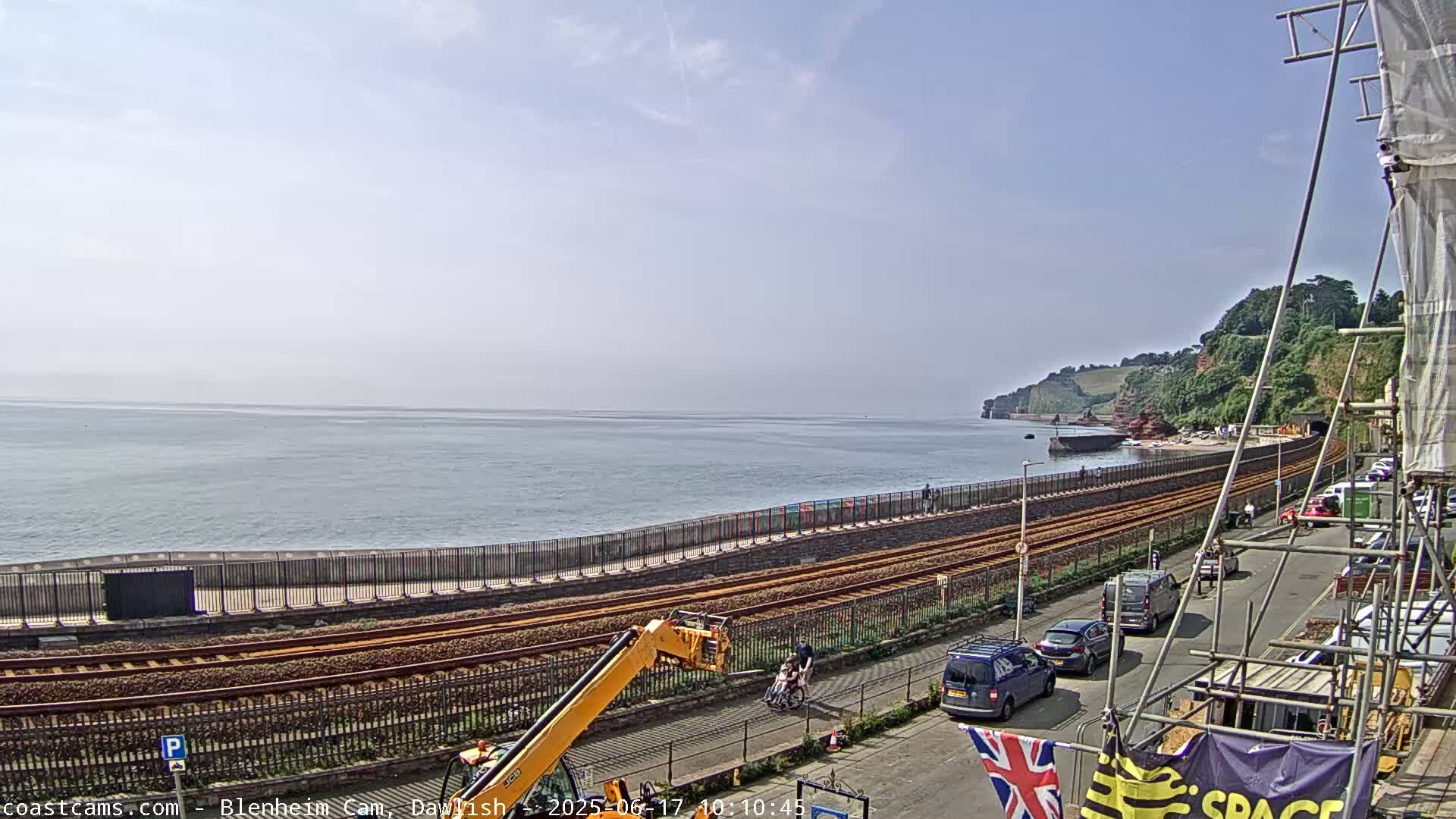 A coastal scene on a partly sunny day shows train tracks running parallel to a seawall and a road with cars parked alongside, overlooking a calm body of water and cliffs in the distance.