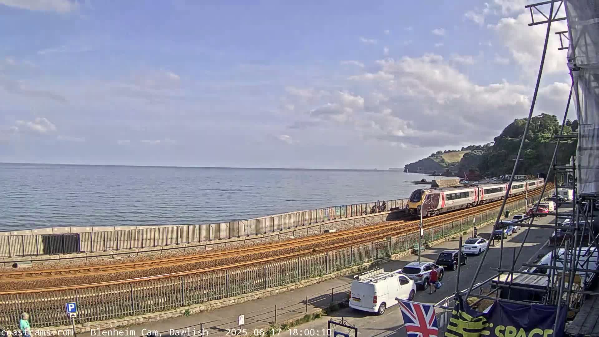 A passenger train travels along a coastal railway line next to a calm sea under a partly cloudy sky.