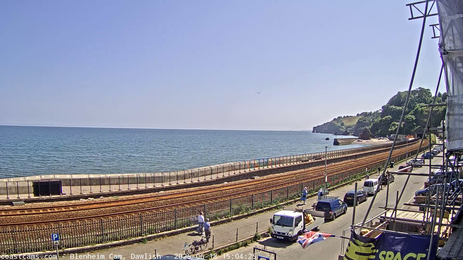 A sunny seaside scene shows railway tracks running alongside a paved road next to a sea wall, with a calm ocean and green cliffs in the distance.