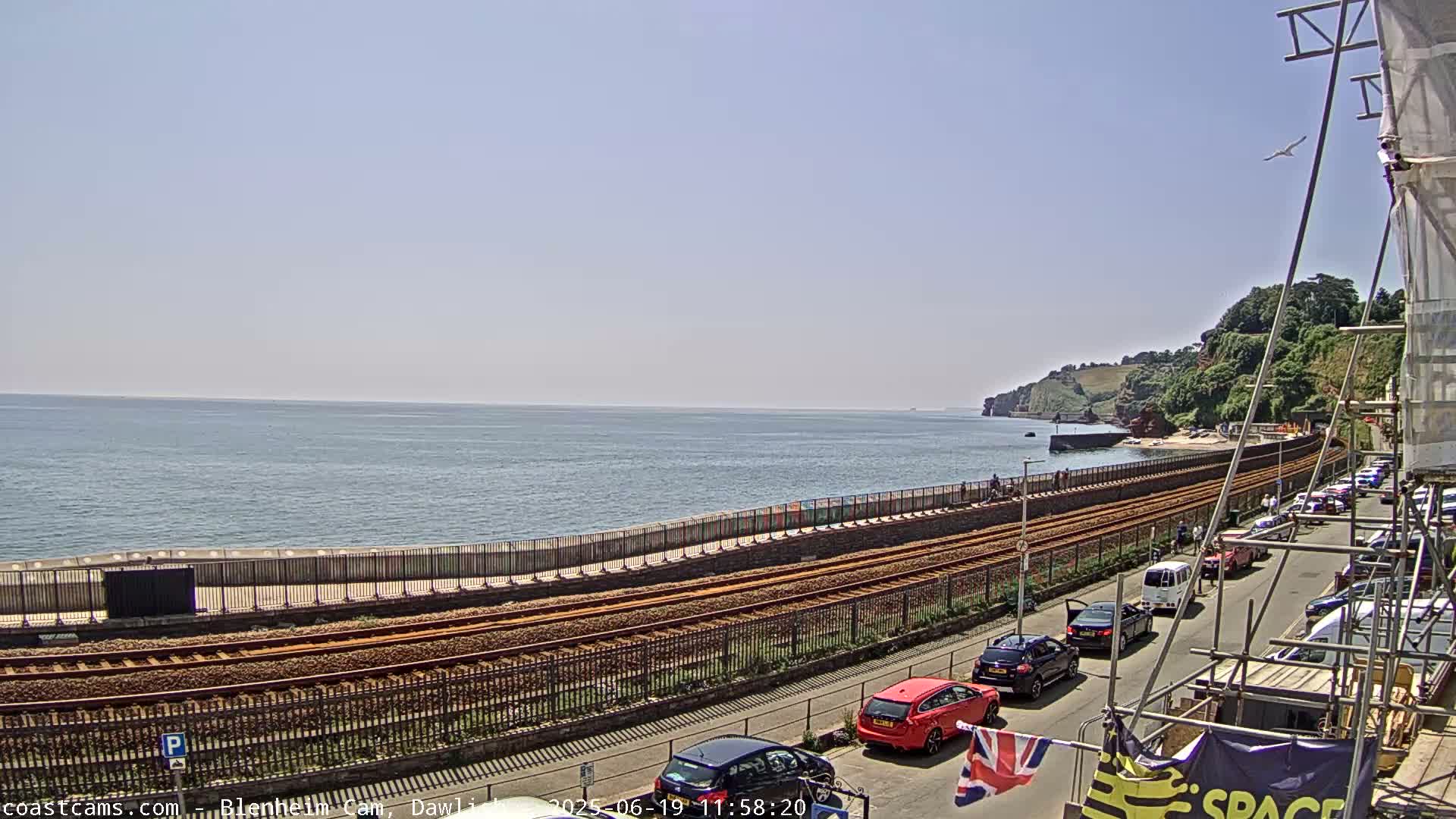 A sunny day at the coast shows a railway track running alongside a sea wall, with cars parked along a road next to it, all overlooking a calm sea and distant cliffs.