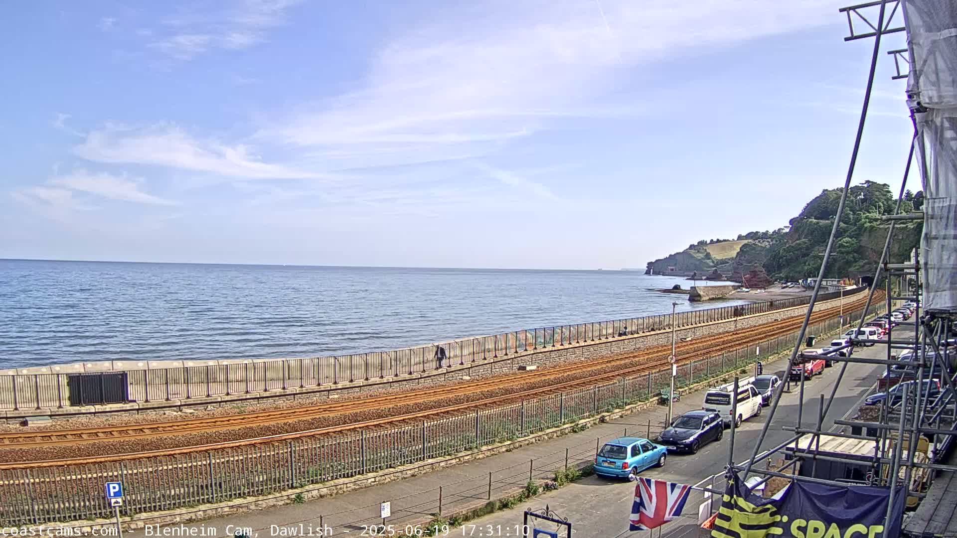 A seaside view on a sunny day shows train tracks running alongside a seawall, with parked cars lining a street beyond.