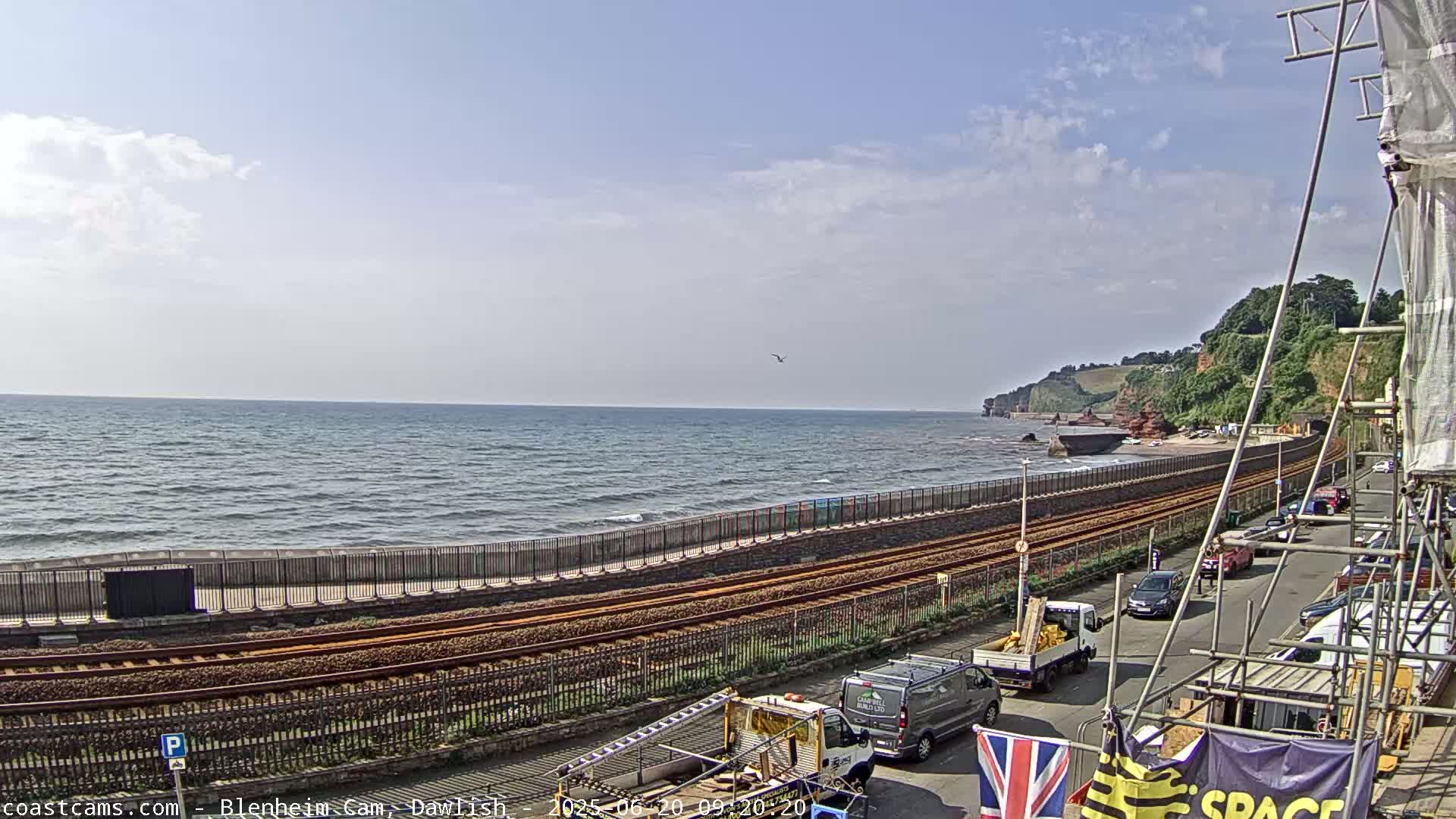 A partly cloudy day shows a coastal scene with a train track running along a seawall next to a beach, with several vehicles parked along the road beyond.