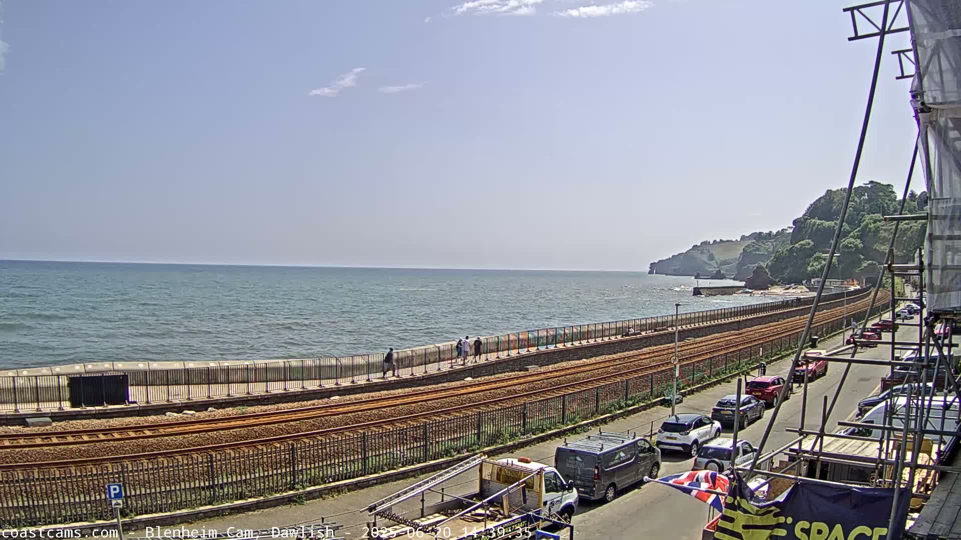 A coastal scene on a sunny day shows a railway line running alongside a seawall, with people walking along the wall and cars parked on the street beyond.