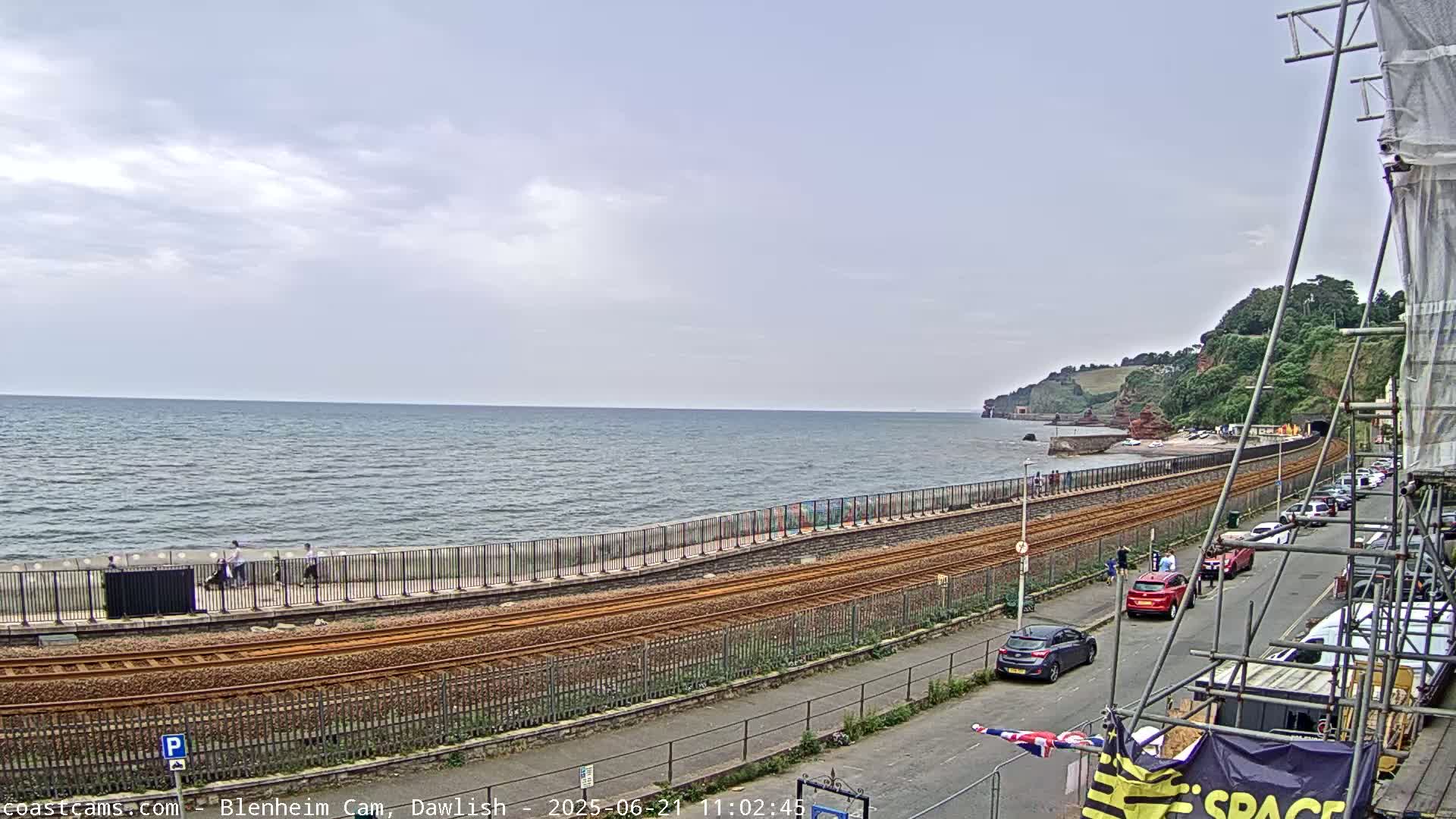 A seaside view shows a train track running alongside a seawall, with cars parked on a street above and a calm sea under a partly cloudy sky.