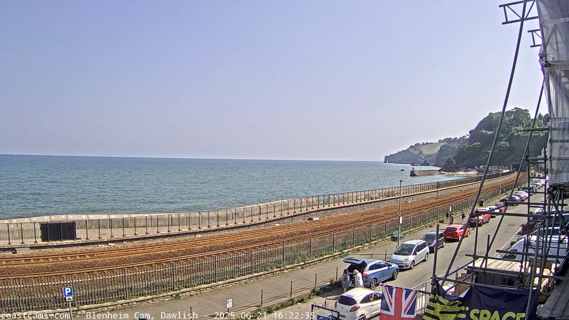 A seaside railway track runs alongside a road lined with parked cars, with a calm sea and cliffs in the background under a clear sky.