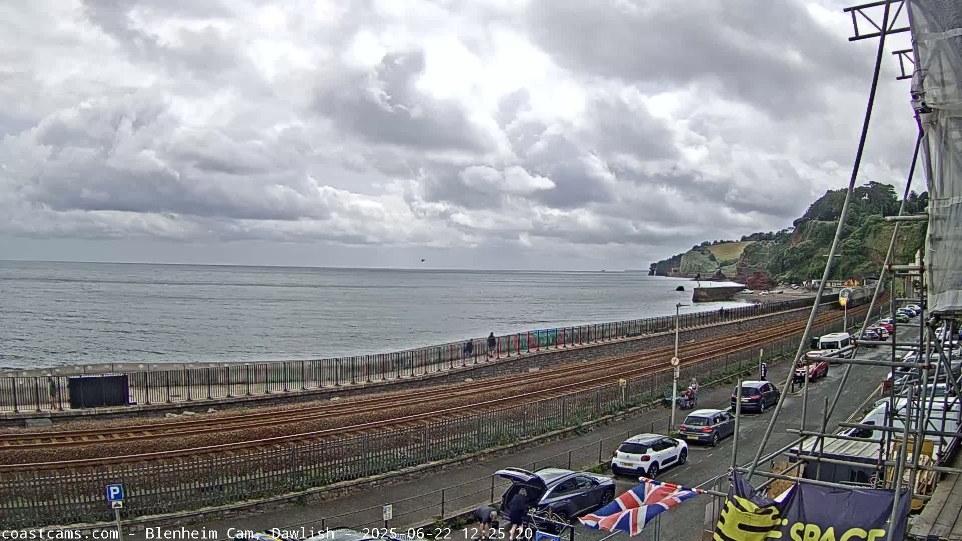 A coastal scene under a cloudy sky shows a train track running alongside a seawall, with cars parked along a street above, and a calm sea extending to a distant, tree-covered headland.