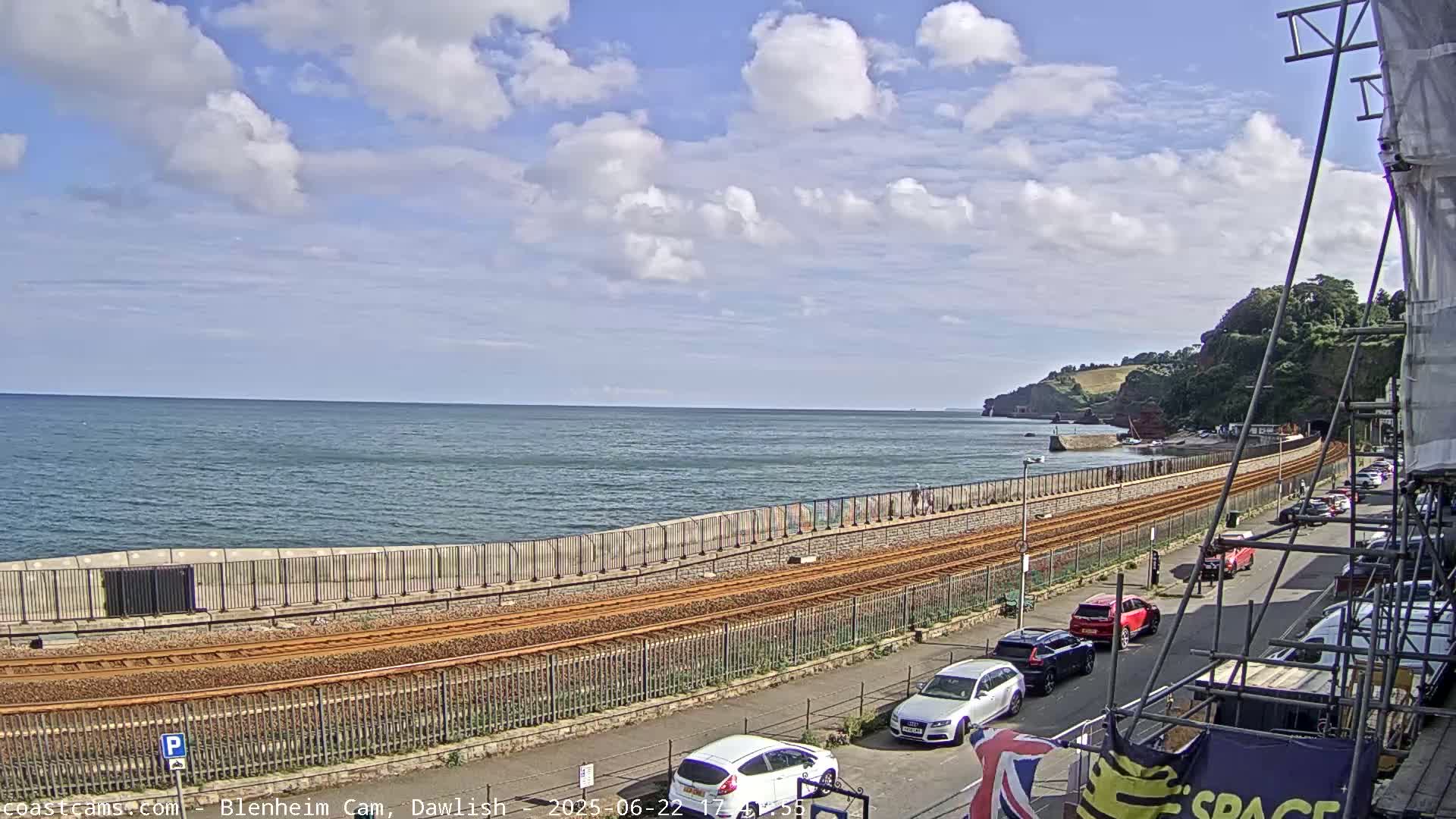 A partly cloudy day shows a coastal scene with train tracks running alongside a seawall, cars parked on a street nearby, and a calm sea in the background.