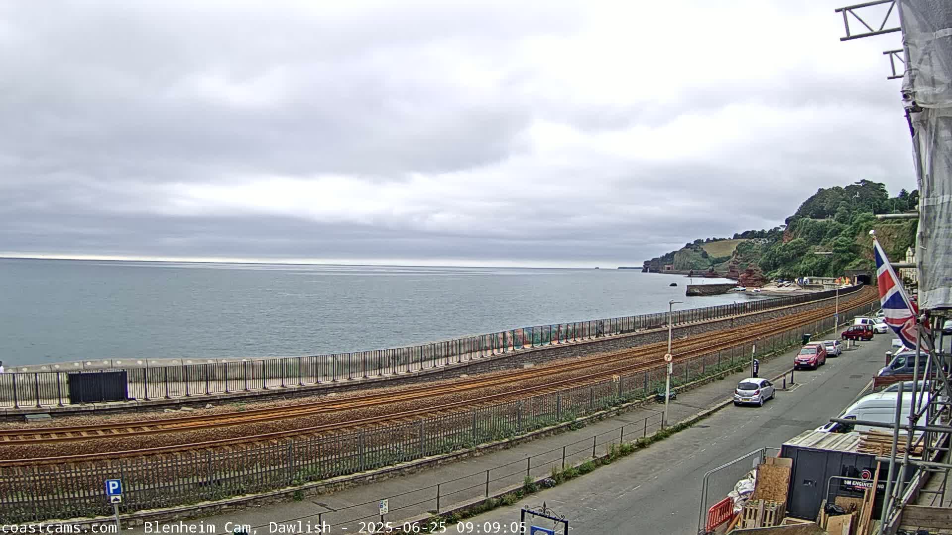 A coastal railway line runs alongside a sea wall, with cars parked on a street next to it, under an overcast sky.