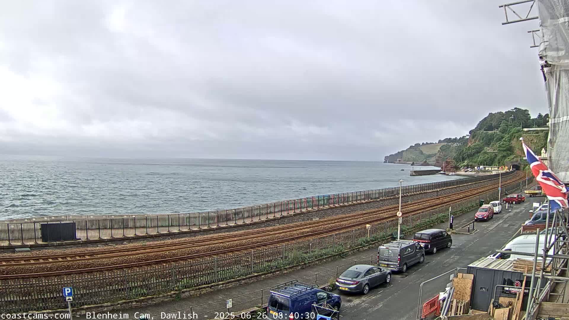 A coastal railway line runs alongside a sea wall, with overcast skies and a calm sea in the background, and several parked cars visible on a nearby road.