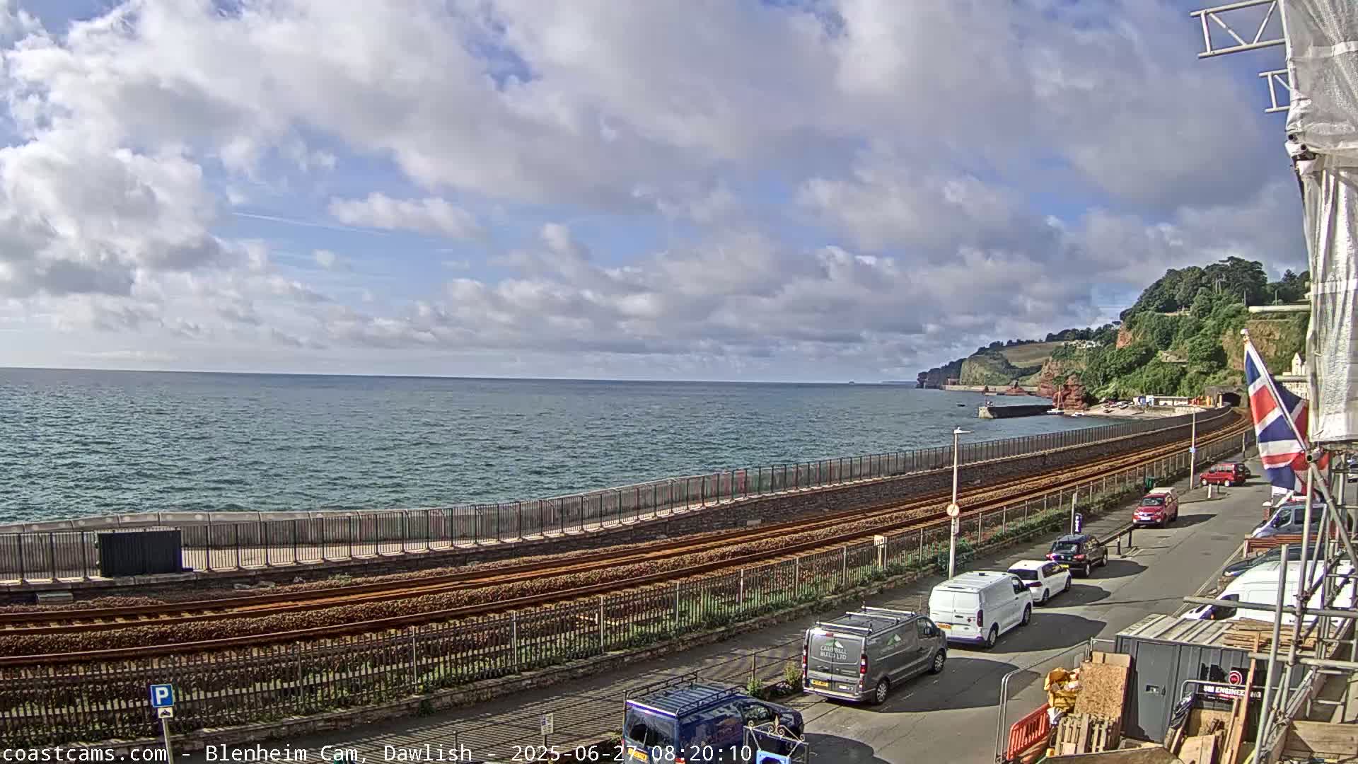 A partly cloudy day shows a coastal scene with a railway line running along a seawall next to a road with parked cars, overlooking a calm sea and distant cliffs.