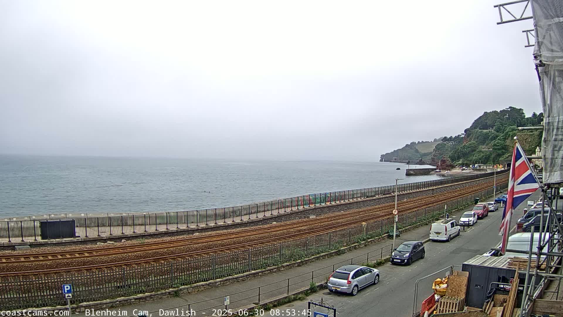 A foggy coastal scene shows railway tracks running alongside a seawall, with a road and parked cars beyond, all overlooking a calm sea and distant cliffs.