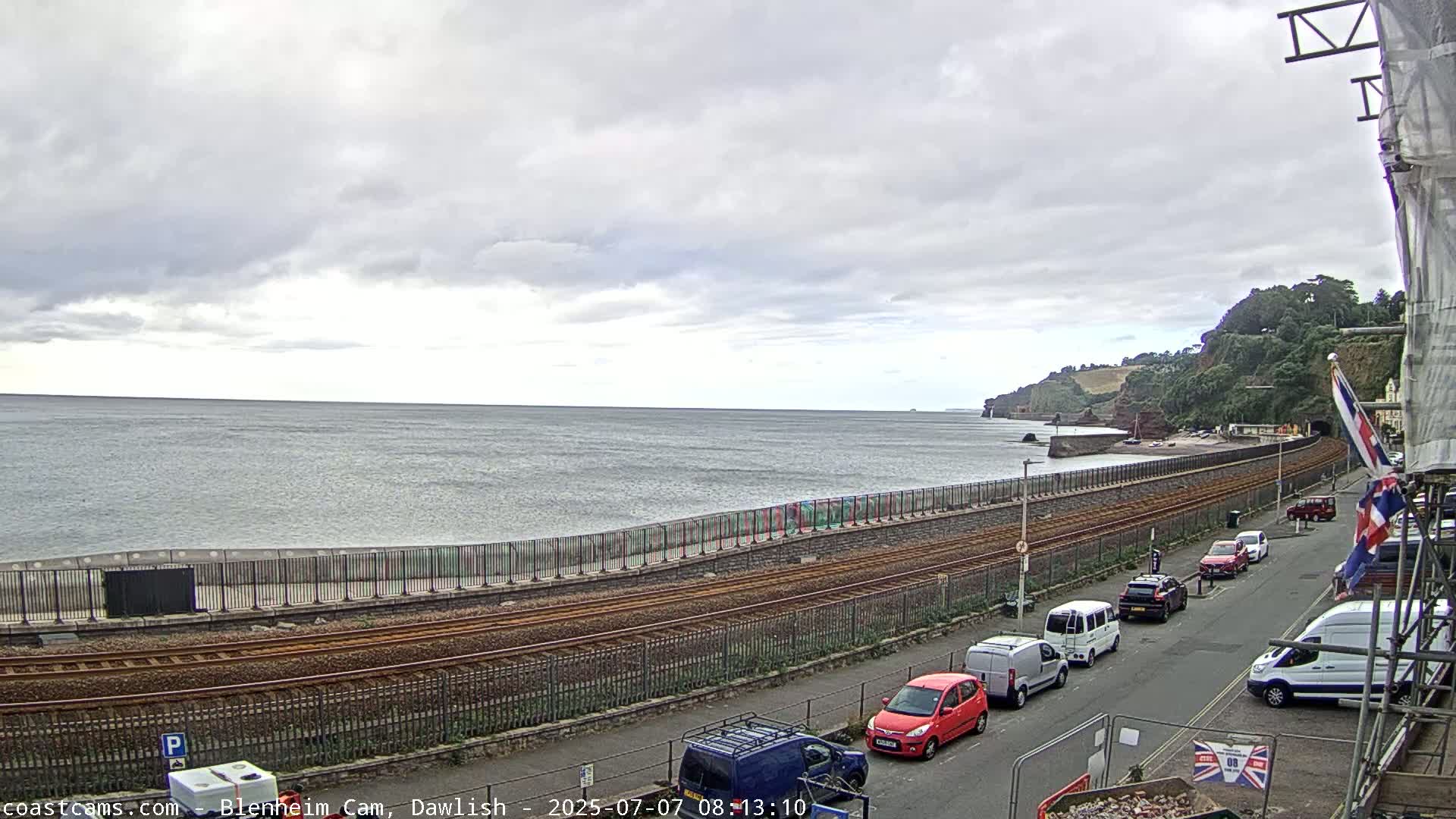 A coastal scene under overcast skies shows railway tracks running alongside a sea wall, with cars parked on a street above, and a calm sea beyond.