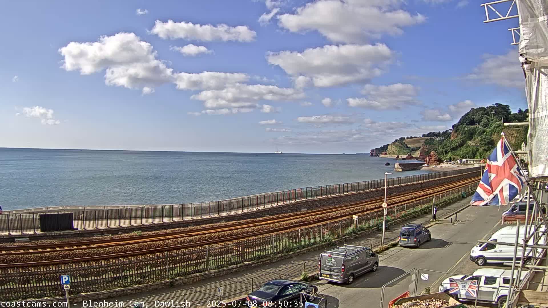A partly cloudy day reveals a coastal scene with railway tracks running alongside a seawall, overlooking a calm ocean and distant cliffs.