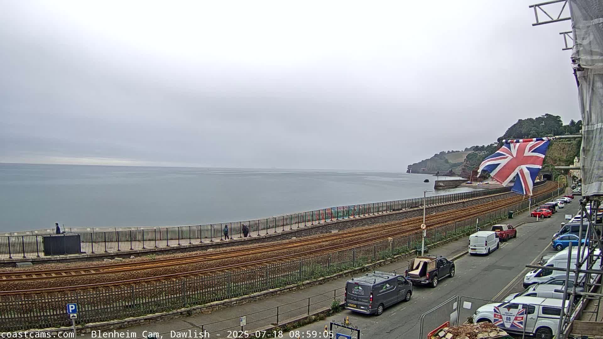 A coastal scene on an overcast day shows a railway line running alongside a seawall, with a Union Jack flag and parked cars visible on a street above.