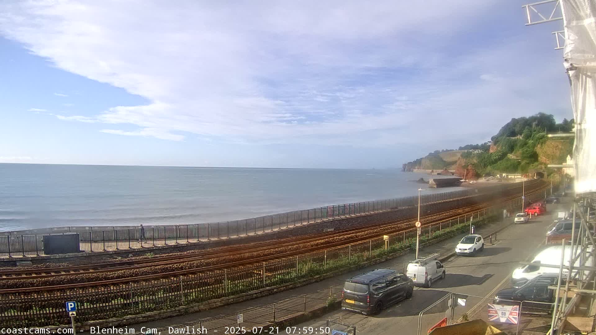 A partly cloudy day overlooks a calm ocean with a shoreline featuring train tracks, a fence, and parked cars.