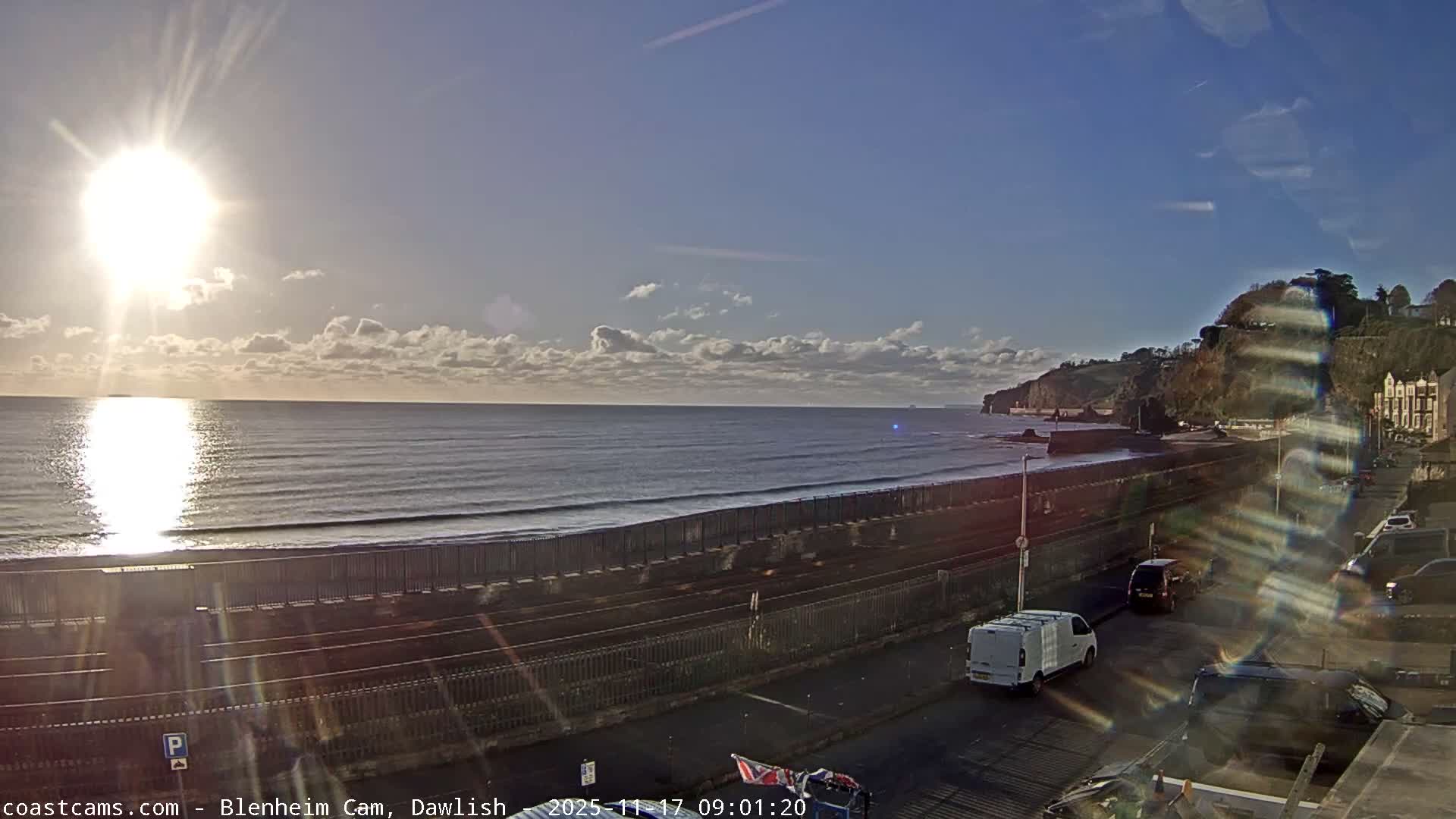 A bright sunny day illuminates a coastal scene featuring a calm sea with reflections, a sandy beach, railway tracks, a road with parked vehicles, and cliffs with some buildings in the distance, under a sky with scattered white clouds.