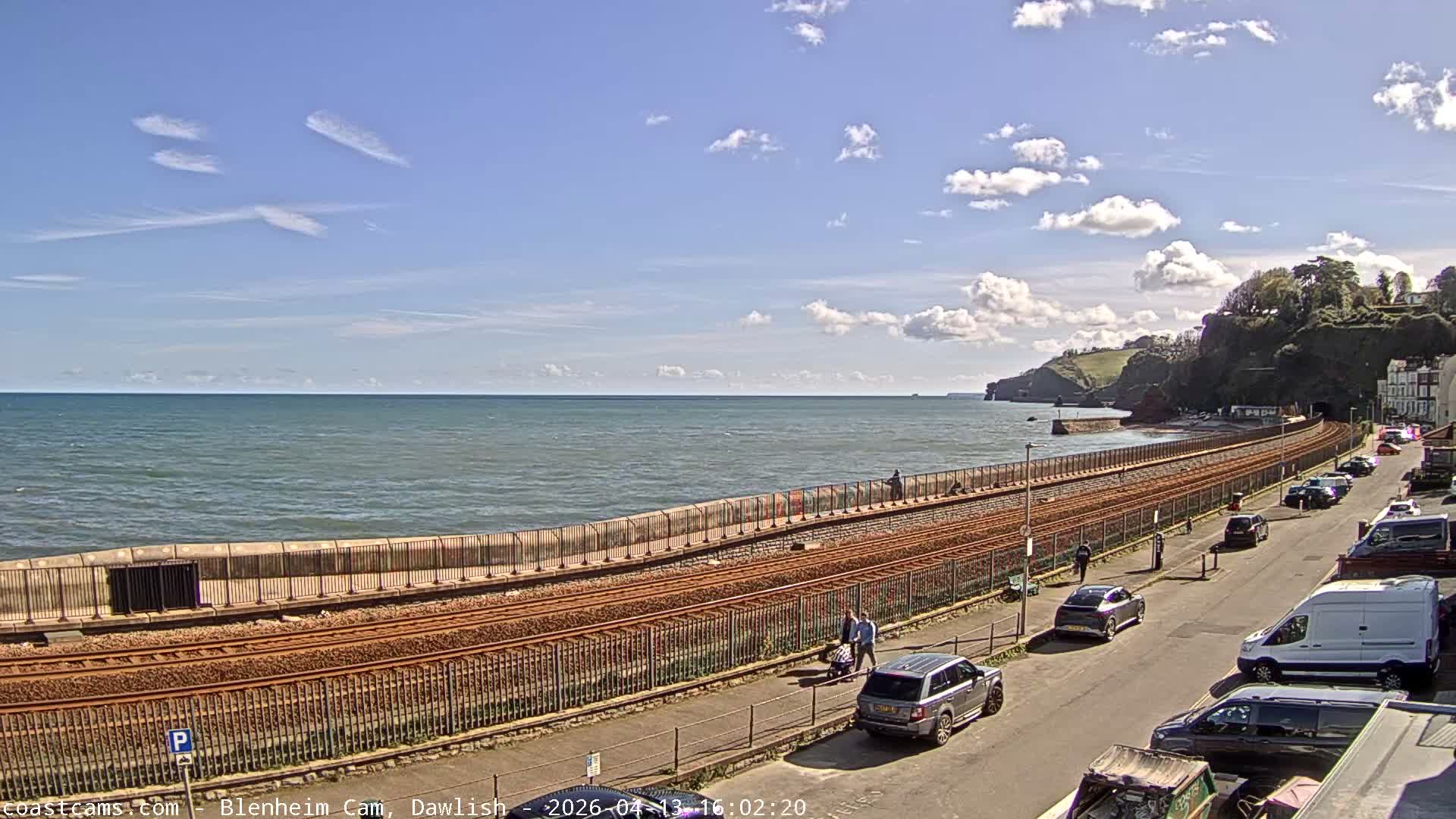 A bright sunny day illuminates a coastal scene featuring a calm sea with reflections, a sandy beach, railway tracks, a road with parked vehicles, and cliffs with some buildings in the distance, under a sky with scattered white clouds.