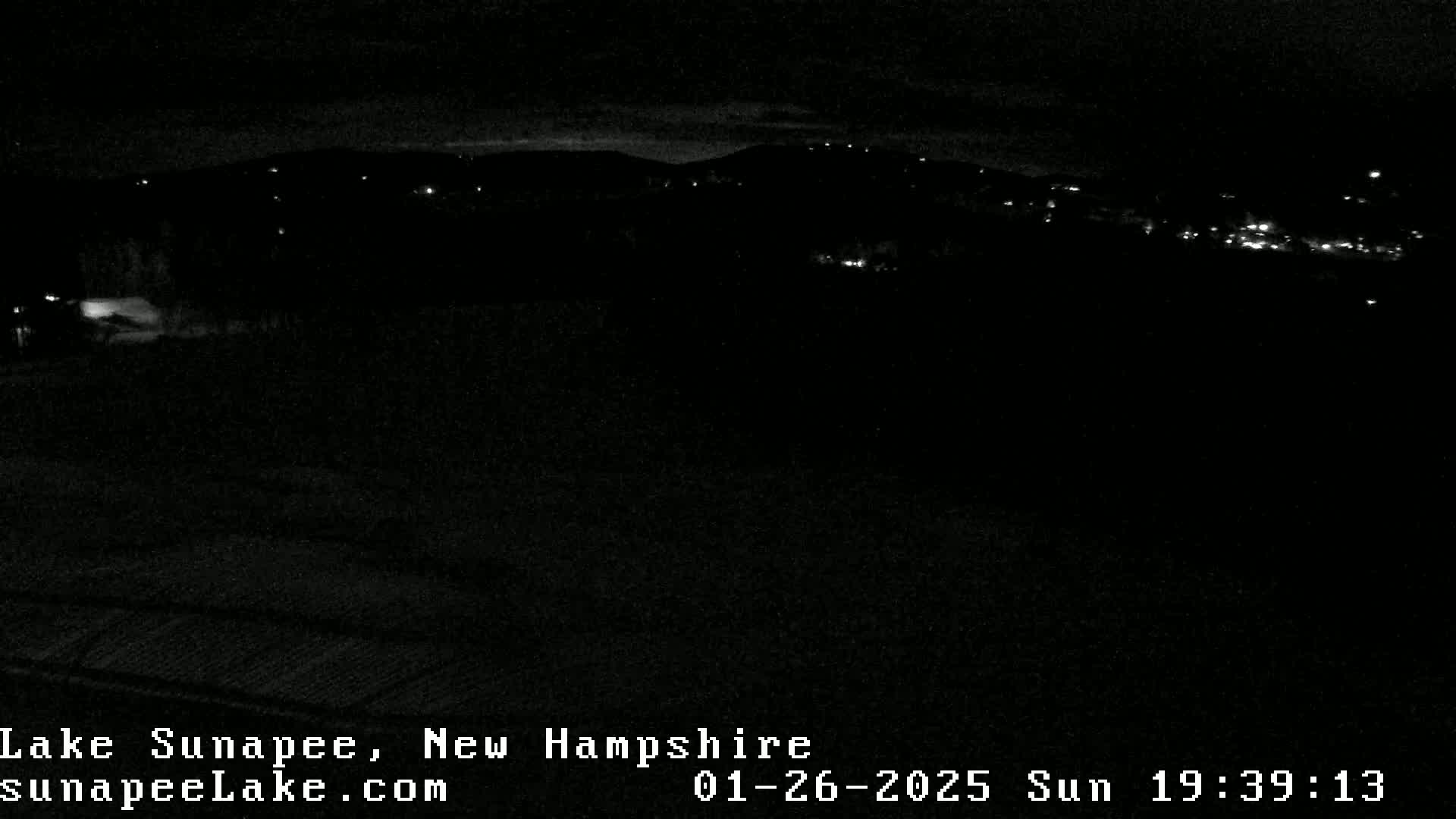 A nighttime, low-light view of a dark landscape with a few distant lights visible on a hillside.
