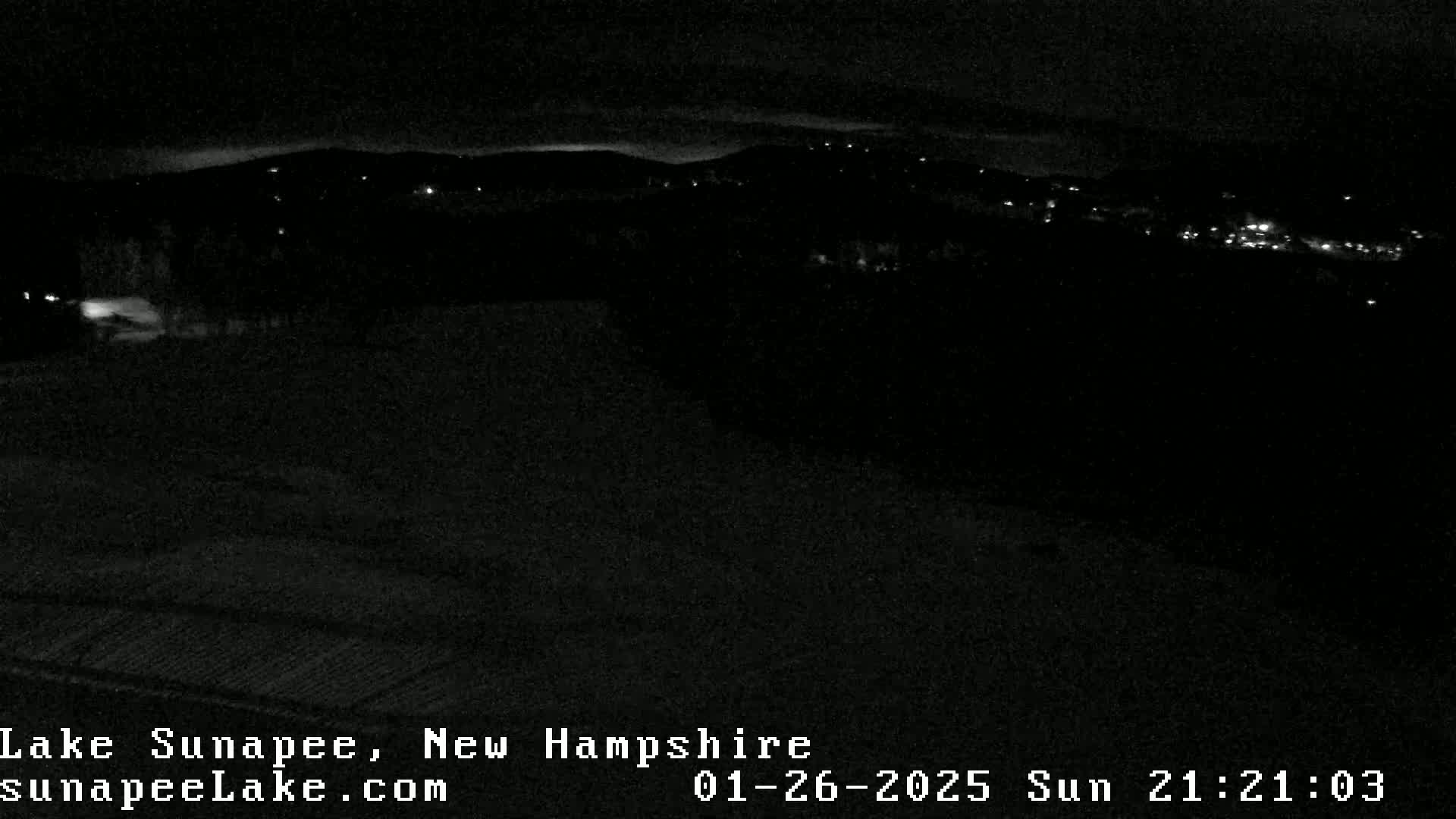 At night, a dark landscape shows a few distant, faint lights from a town or village under a clear, dark sky.