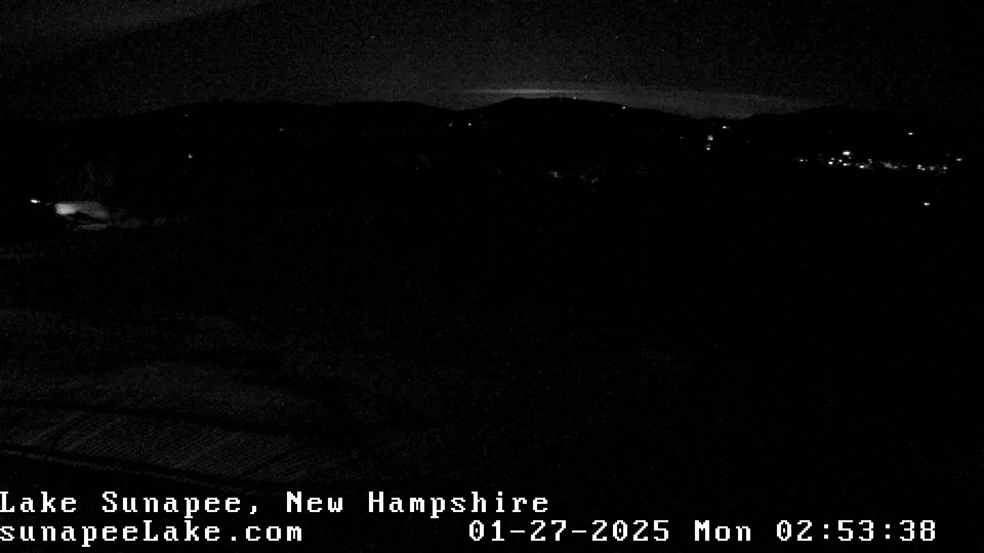 A nighttime, low-light image shows a dark landscape with a few distant lights, possibly a town or village, under a clear sky.