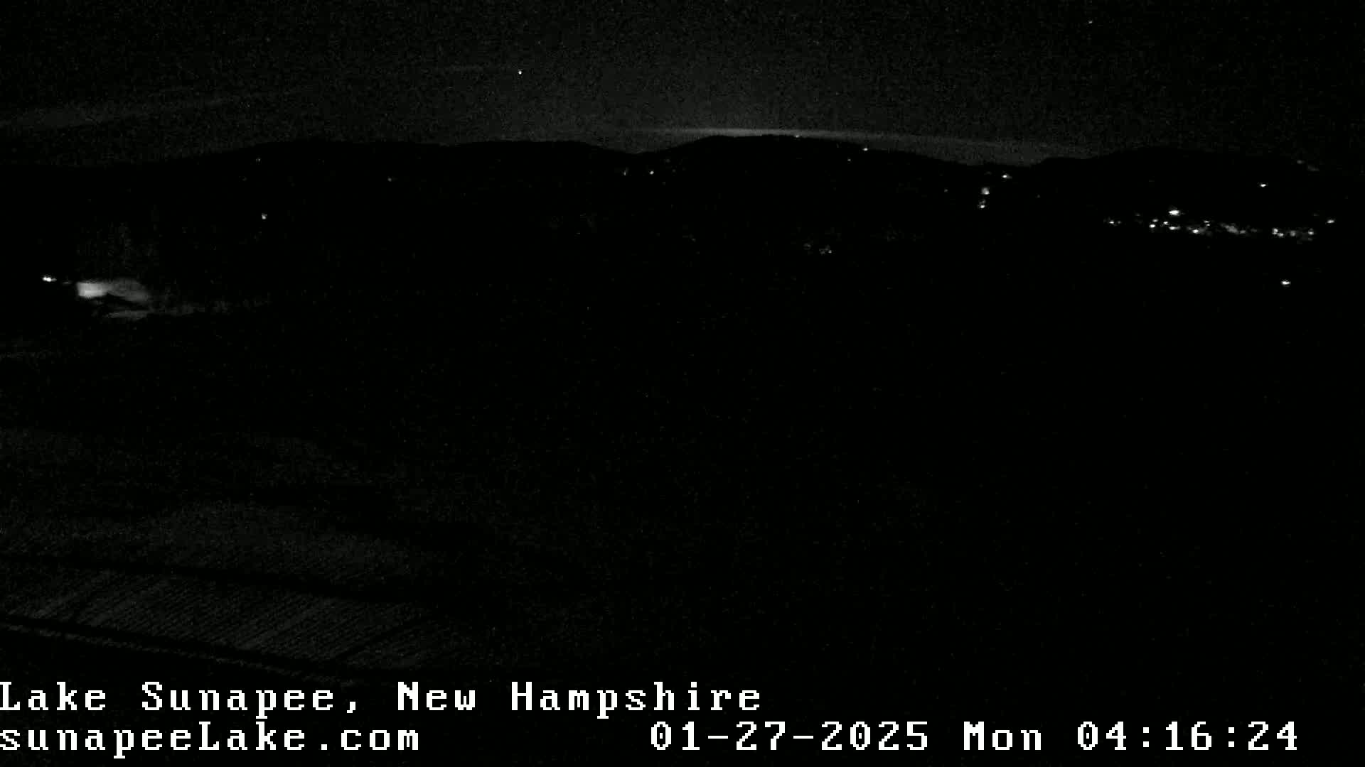 A nighttime, low-light view shows a dark landscape with a few distant lights, possibly a town or village, under a clear, dark sky.