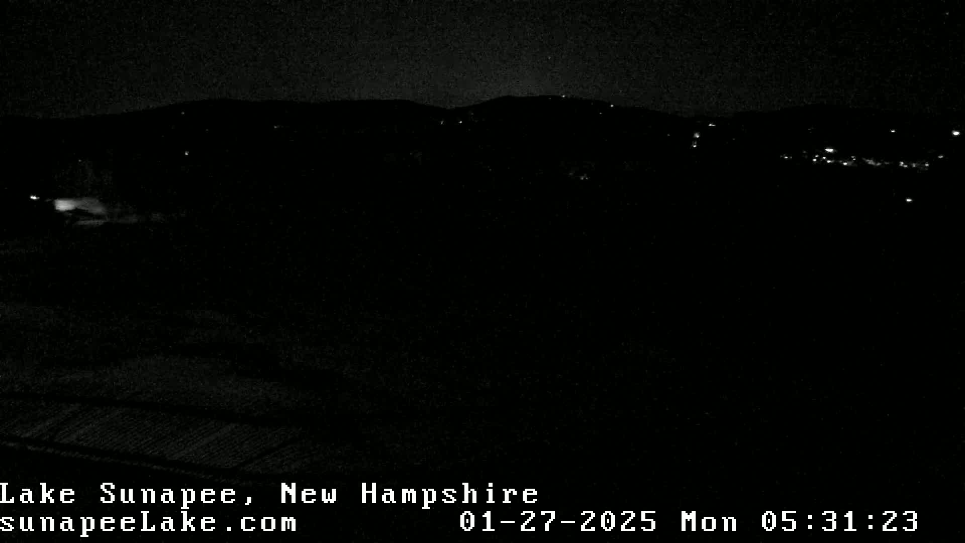 A nighttime, low-light view shows a dark landscape with a few distant lights, possibly a town or village, under a clear, dark sky.