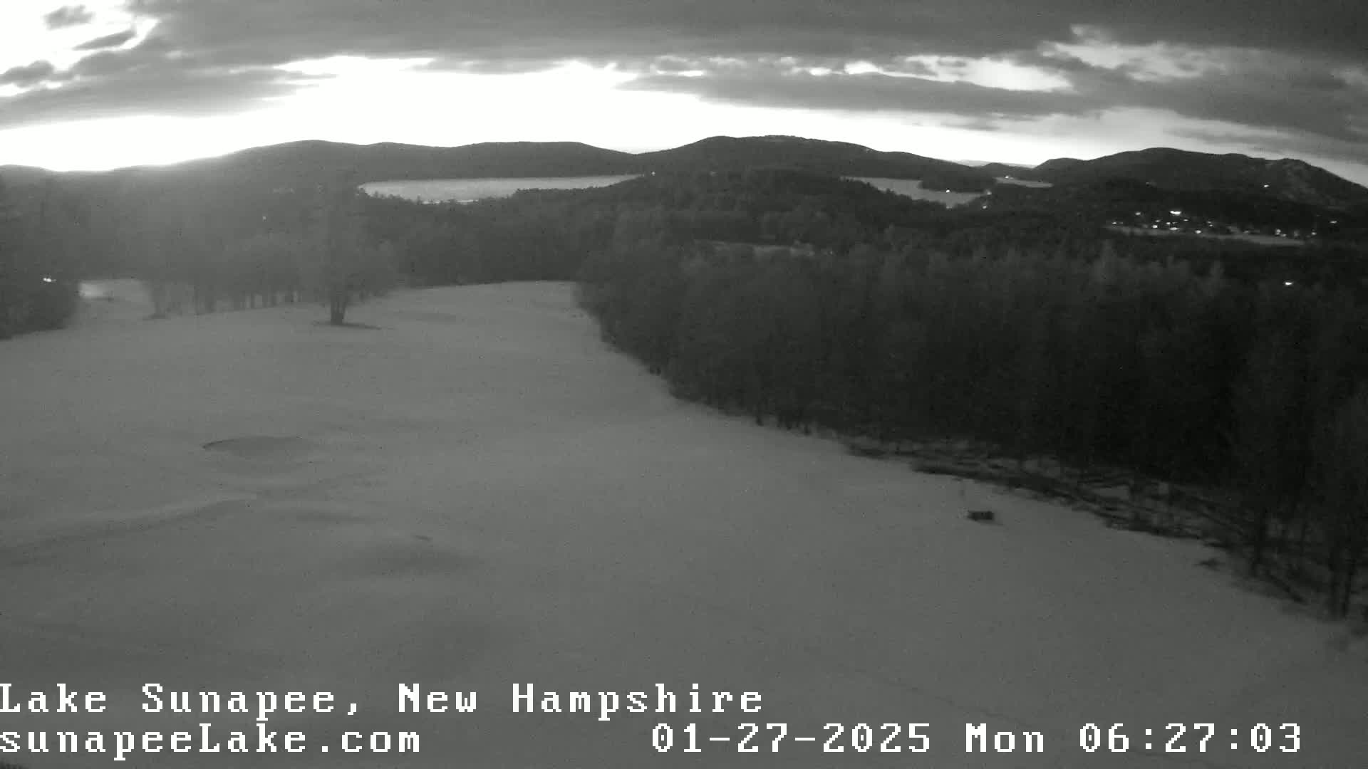 A snow-covered landscape at dawn, showing a golf course, trees, and a lake in the distance under a cloudy sky.