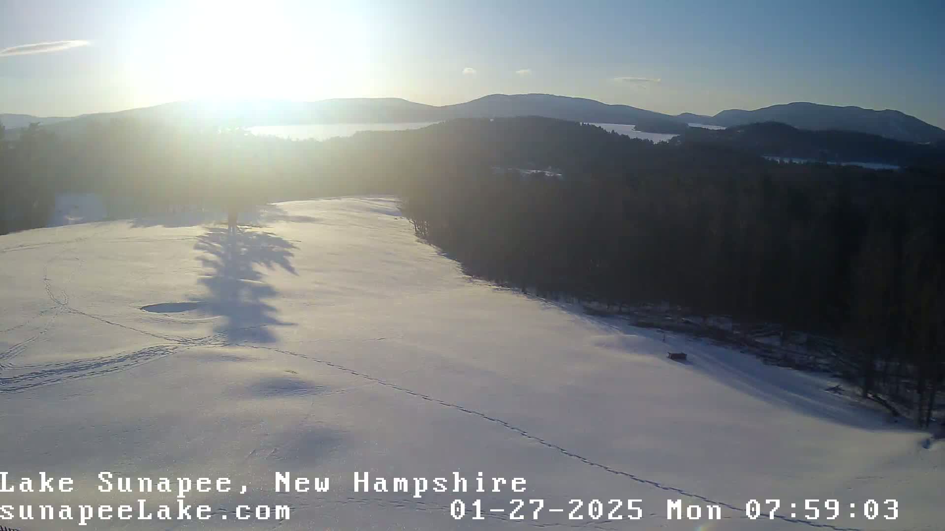 A snow-covered field with animal tracks stretches toward a sunlit lake and distant mountains under a clear, bright sky.
