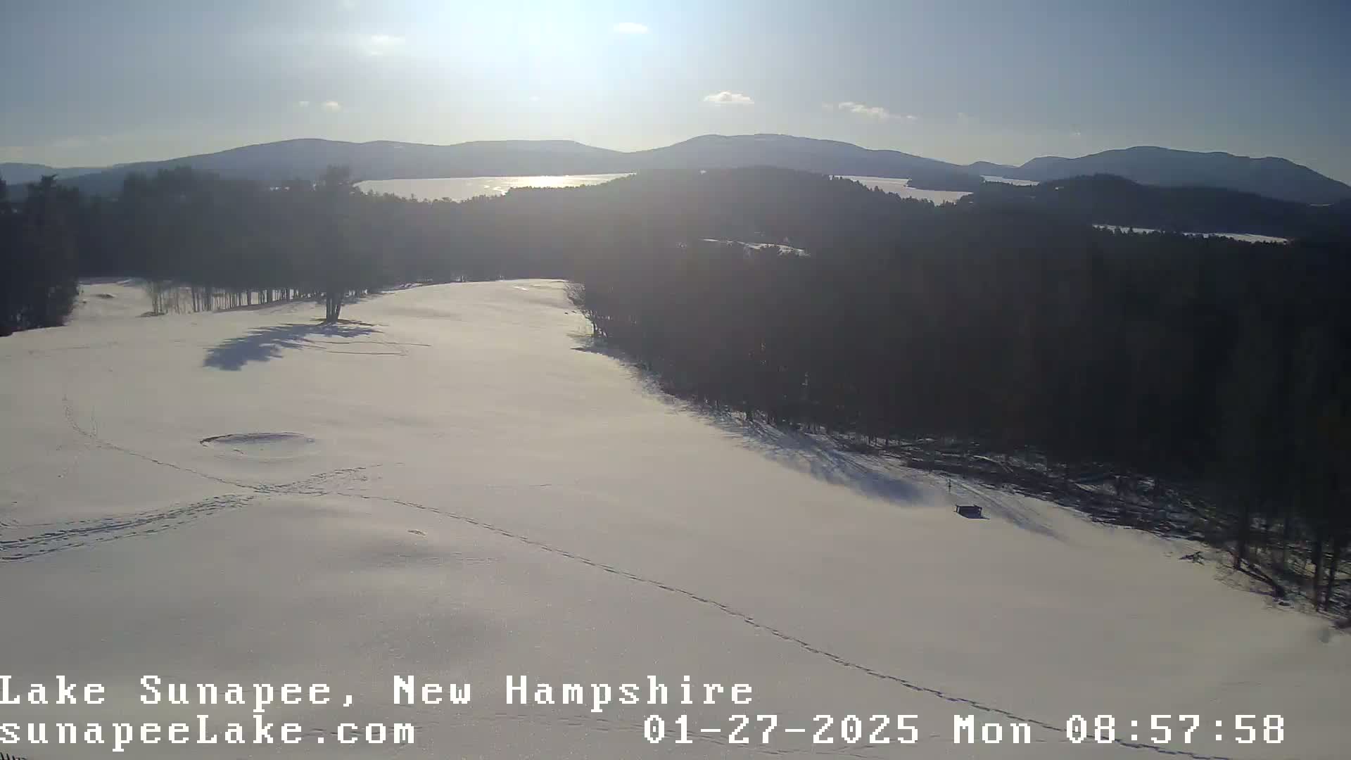 A snow-covered landscape with a distant view of a lake and mountains under a bright, sunny sky.