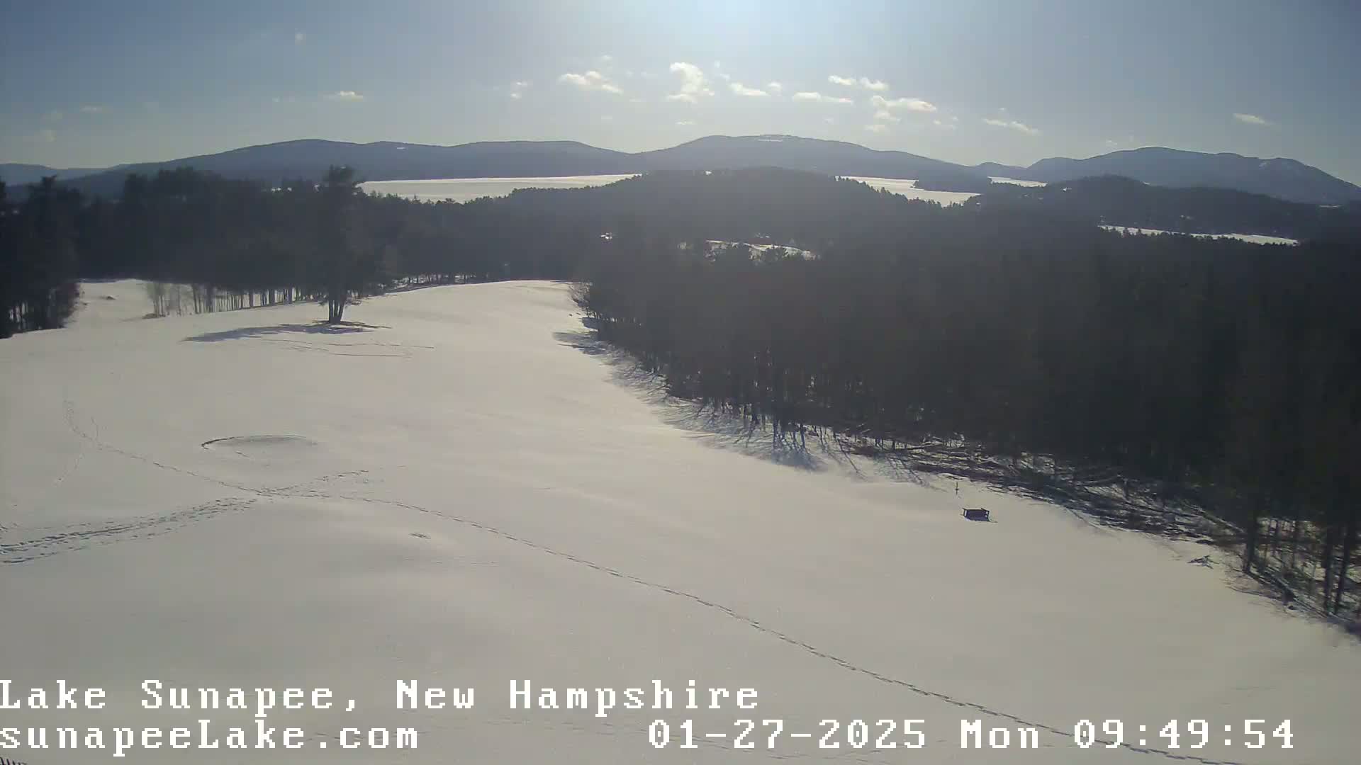 A snow-covered landscape with a lake and distant mountains under a sunny sky.