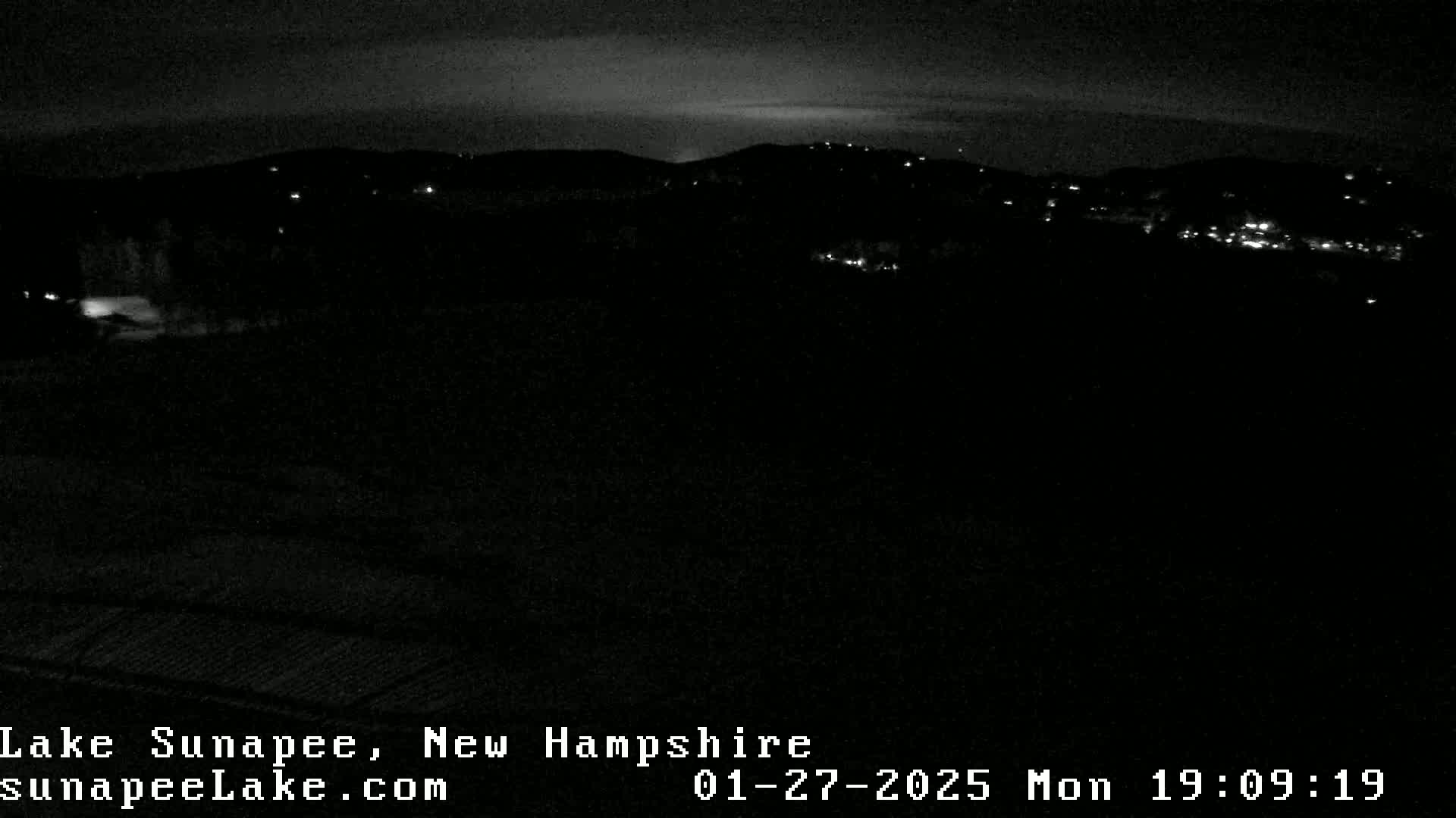 A nighttime long shot of a dark landscape with sparsely scattered lights in the distance under a mostly clear sky.
