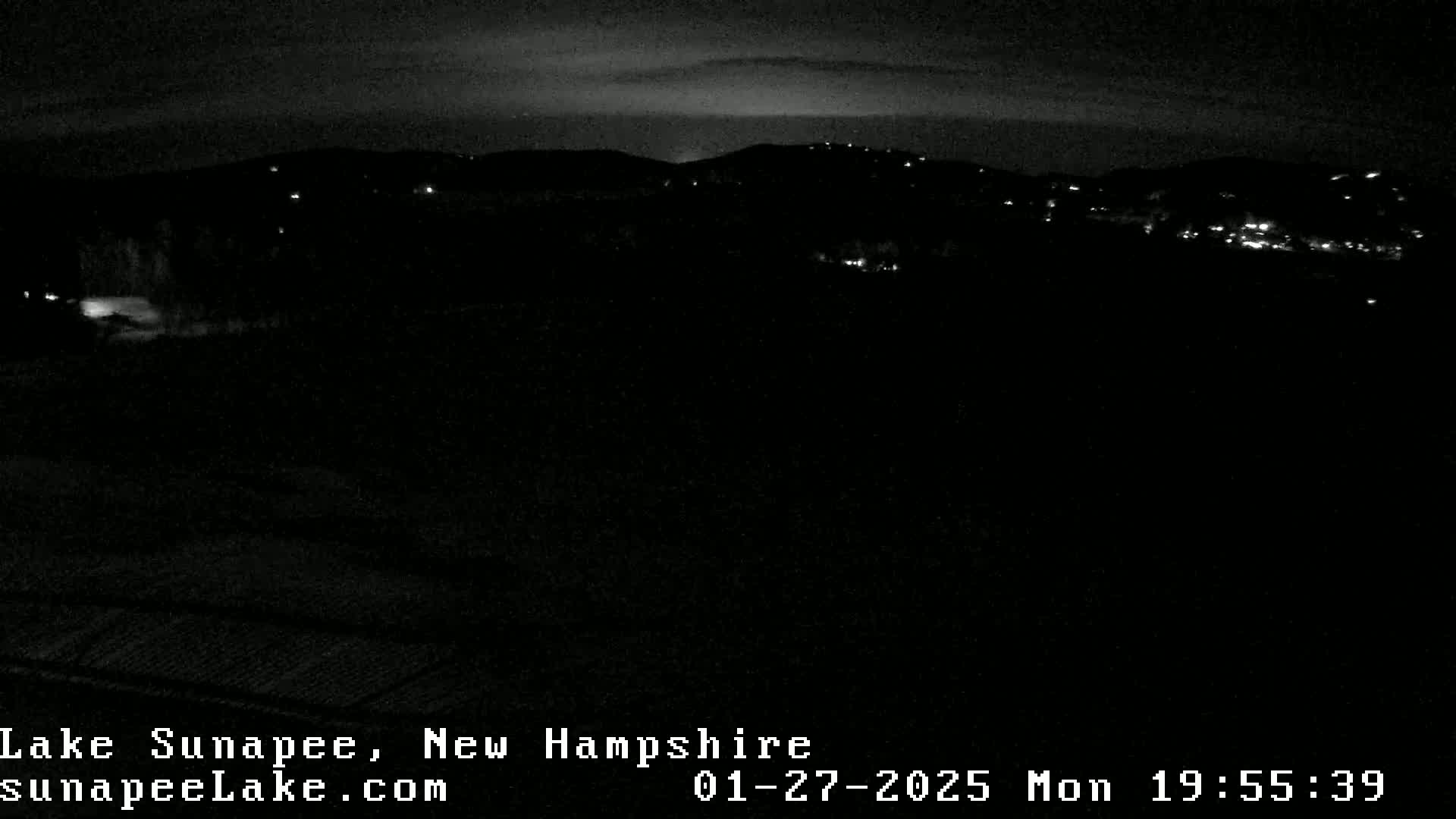 A nighttime view of dimly lit hills and a dark foreground under a mostly clear sky.
