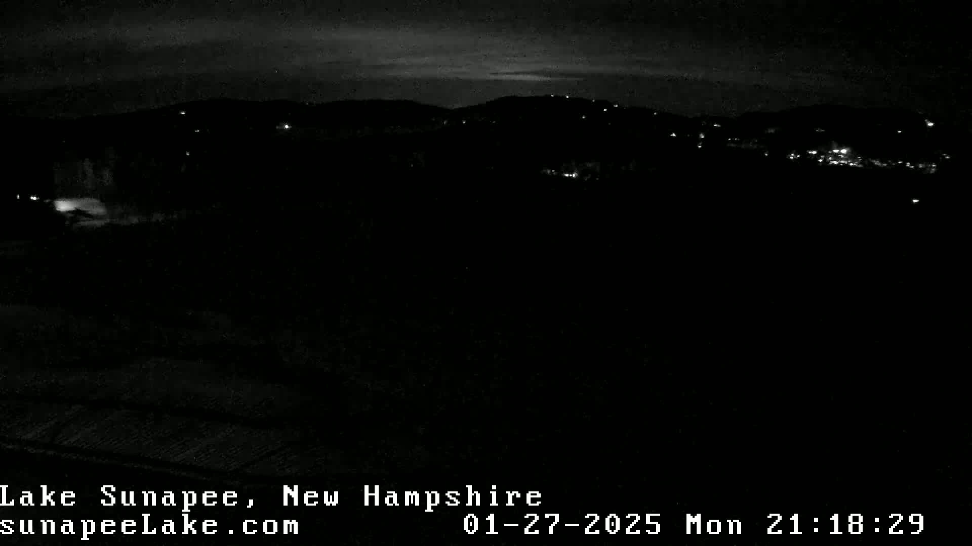 A nighttime, low-light view of a dark landscape with sparsely lit hills in the distance under a mostly clear sky.