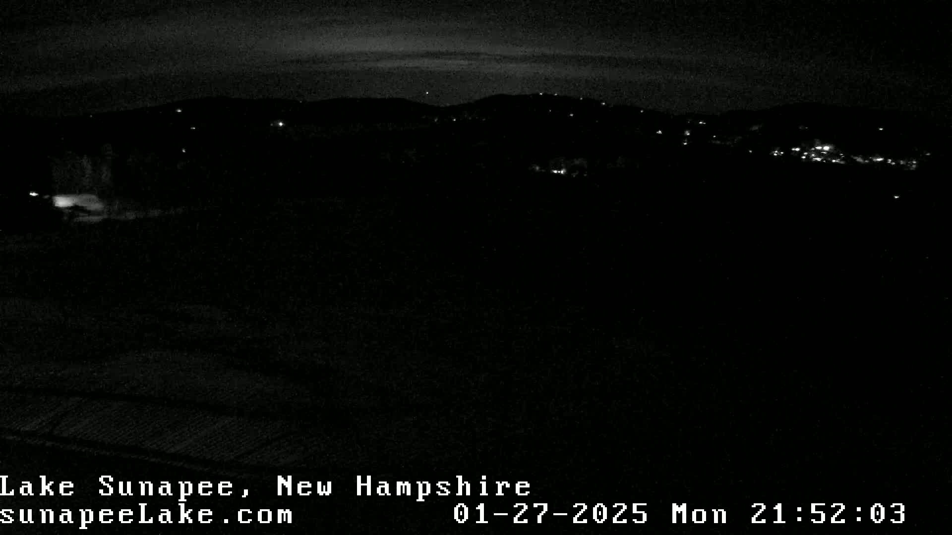 A nighttime, low-light view of a dark landscape with a few distant lights visible under a mostly clear sky.