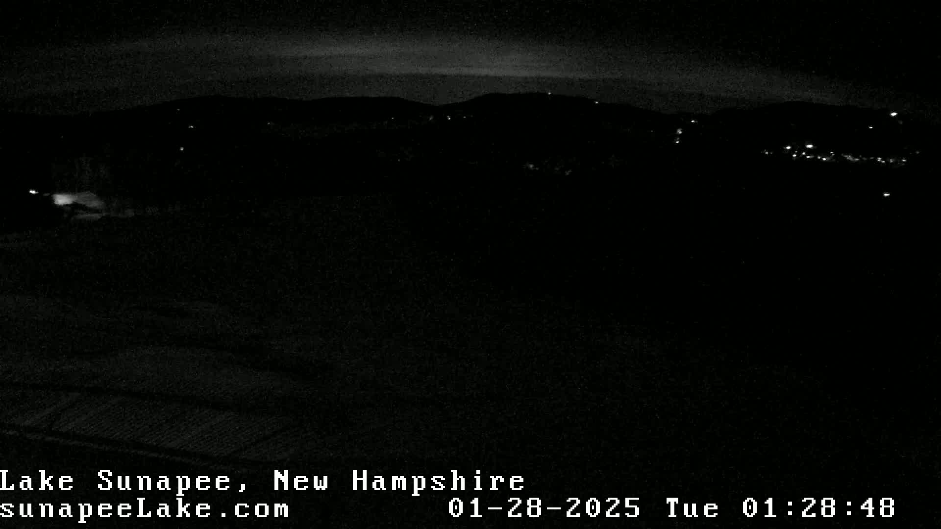 A nighttime, low-light view of dark hills and a few distant lights under a mostly clear sky.