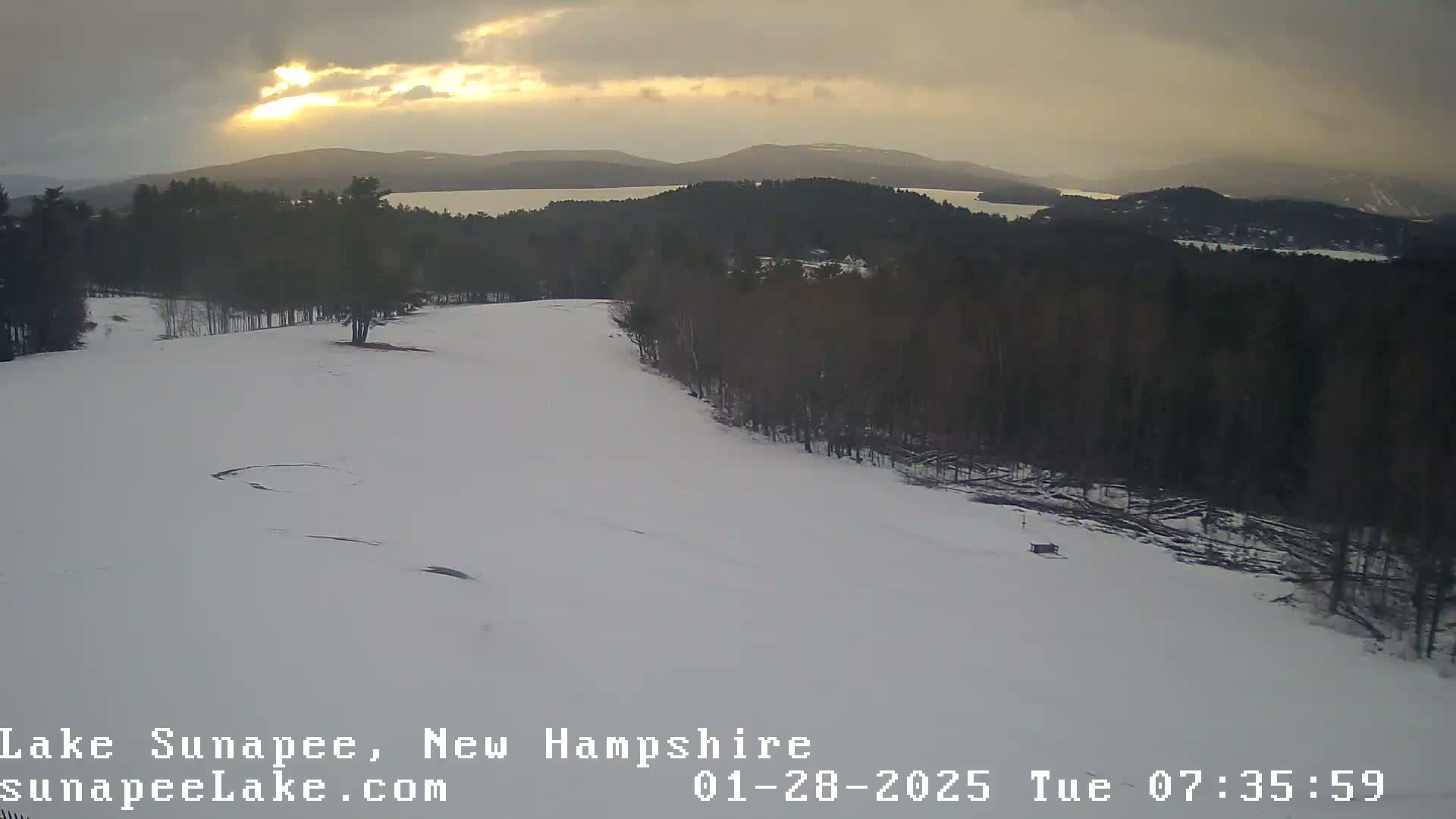 A snow-covered hillside with a forest and a lake in the distance under an overcast sky with a faint sunset.