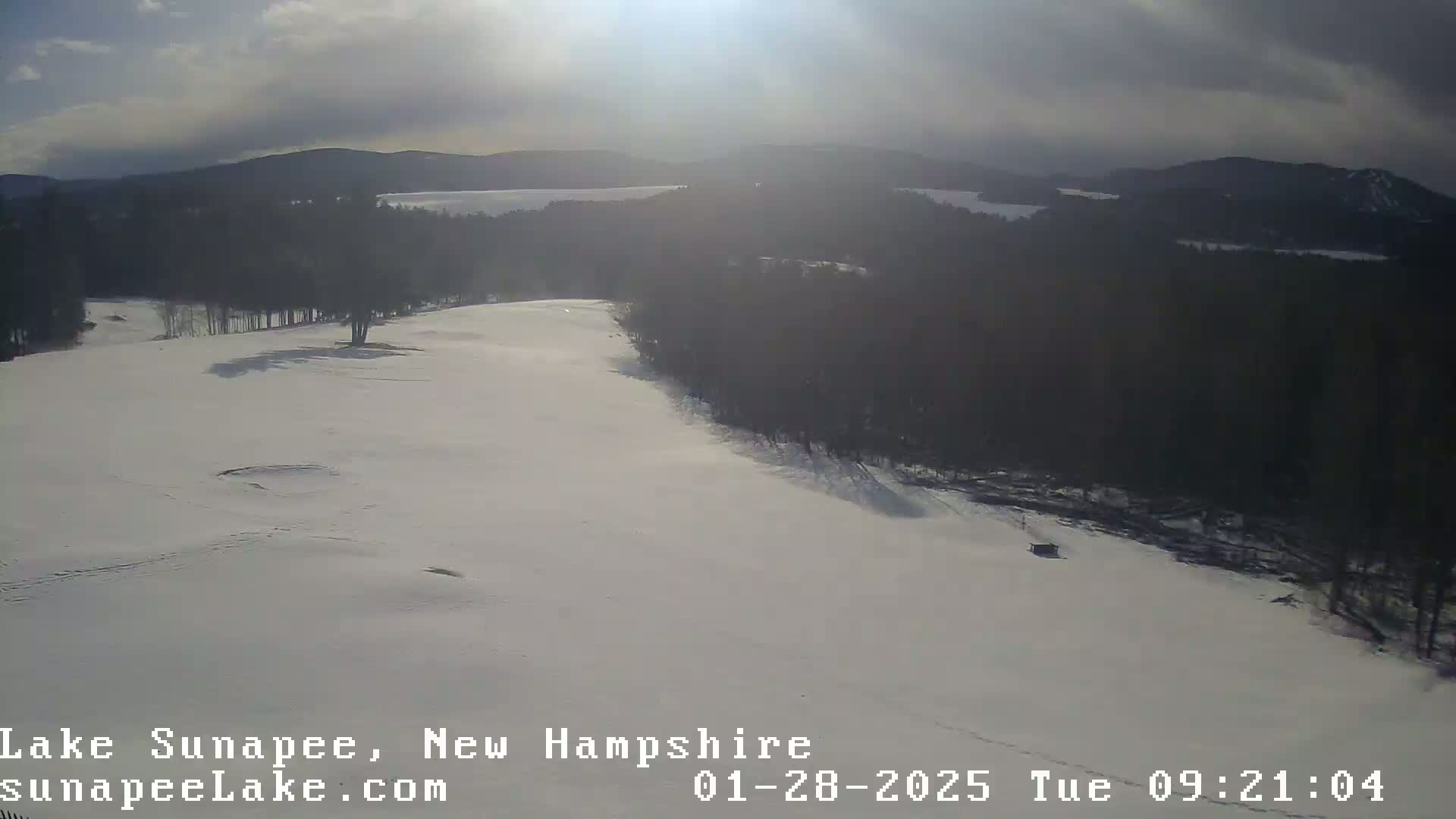 A snow-covered landscape with a partially visible lake in the distance under a partly cloudy sky.