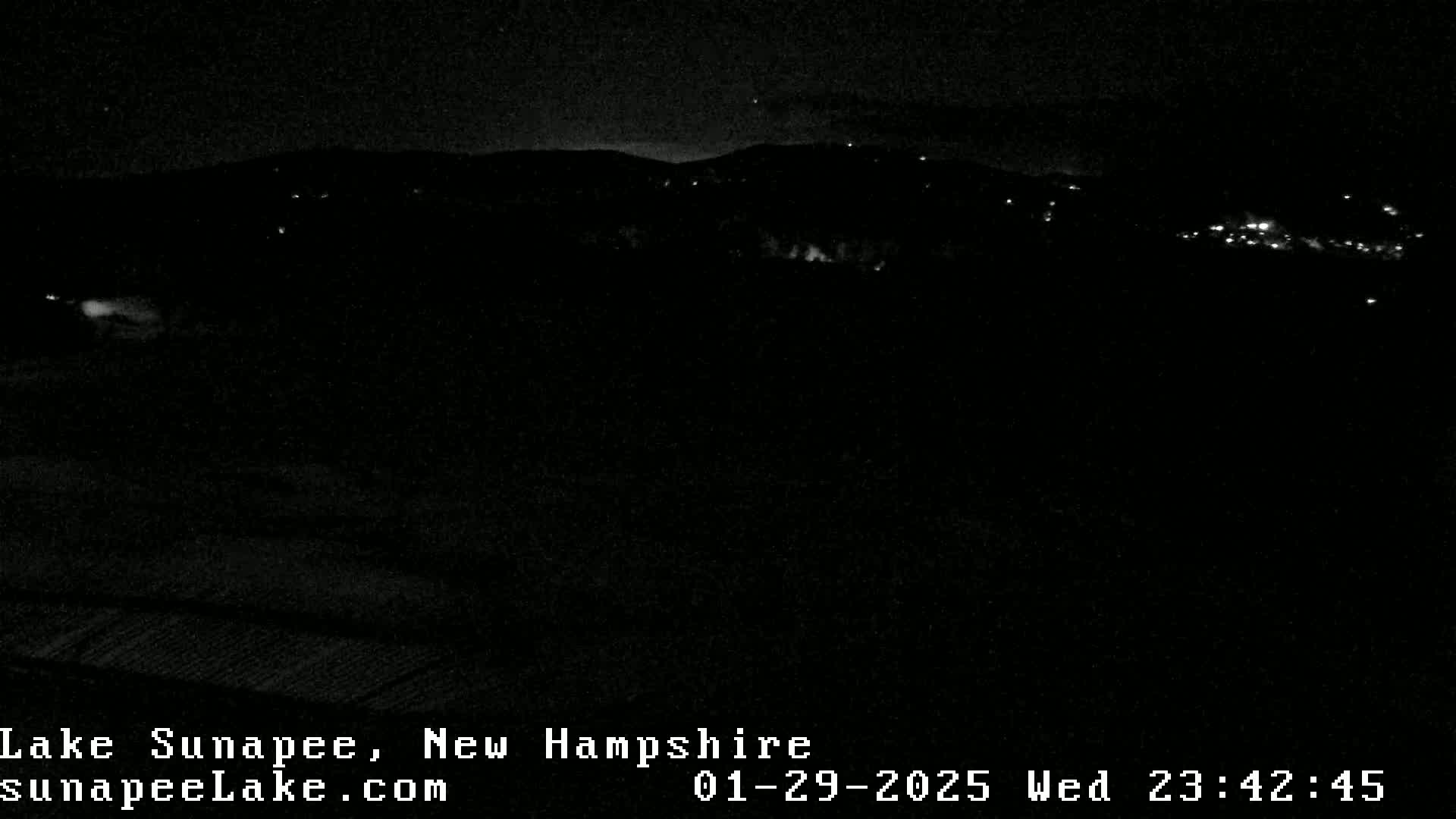 A nighttime, low-light view of a dark landscape with a few distant lights visible, possibly a small town or village, under a clear night sky.