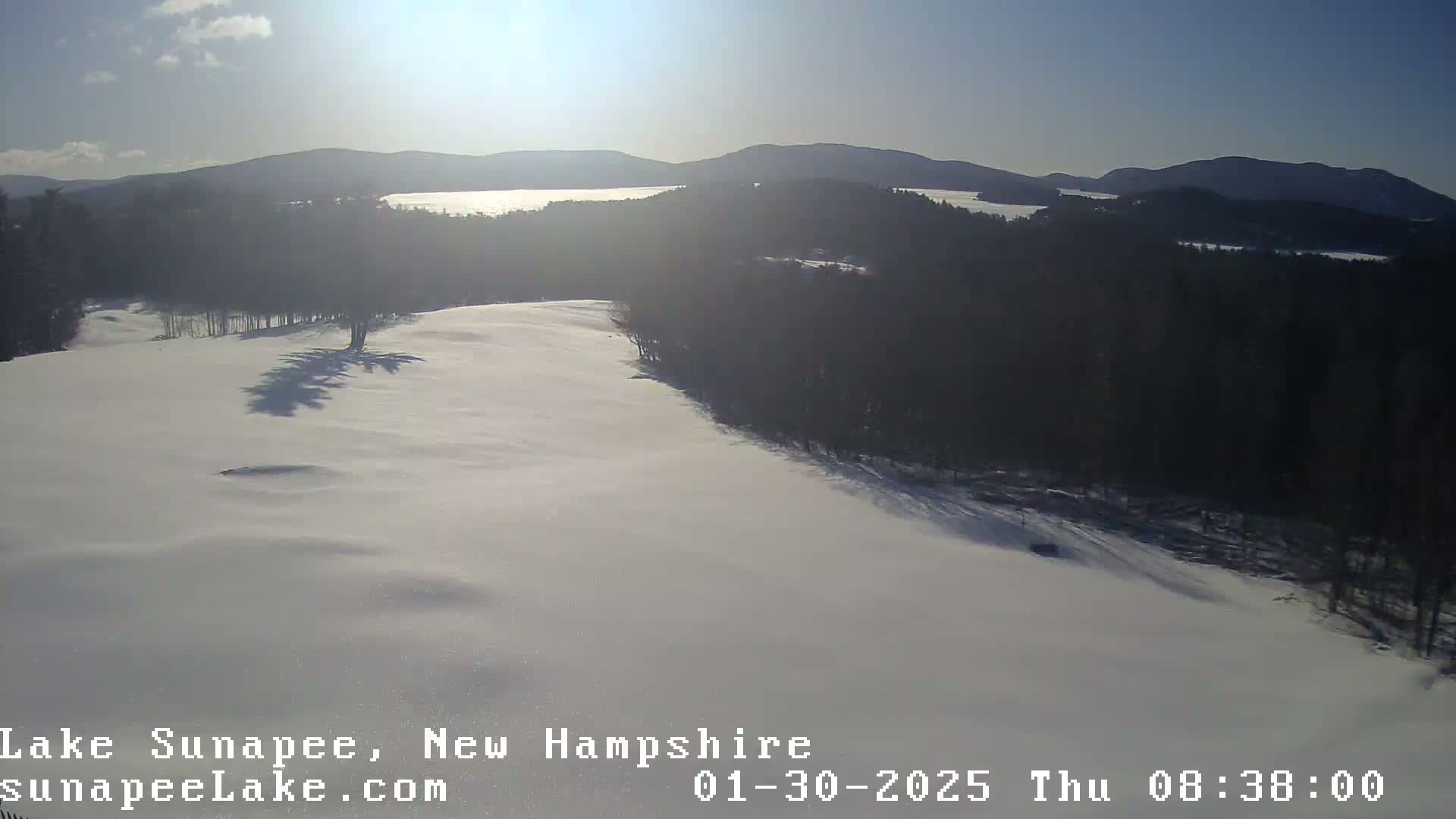 A snow-covered landscape with a distant view of a lake and mountains under a sunny sky.