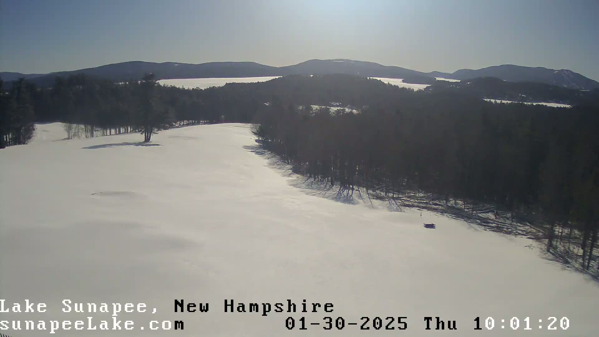 A snow-covered landscape with a dark line of trees separating a large expanse of snow from a distant lake and mountain range under a bright, sunny sky.