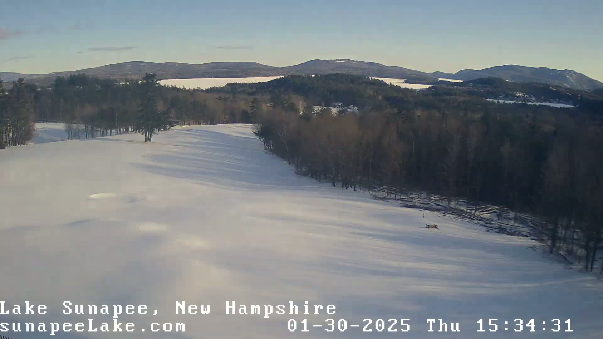 A snow-covered landscape shows a gently sloping hill leading down to a wooded area, with mountains and a lake visible in the distance under a clear sky.