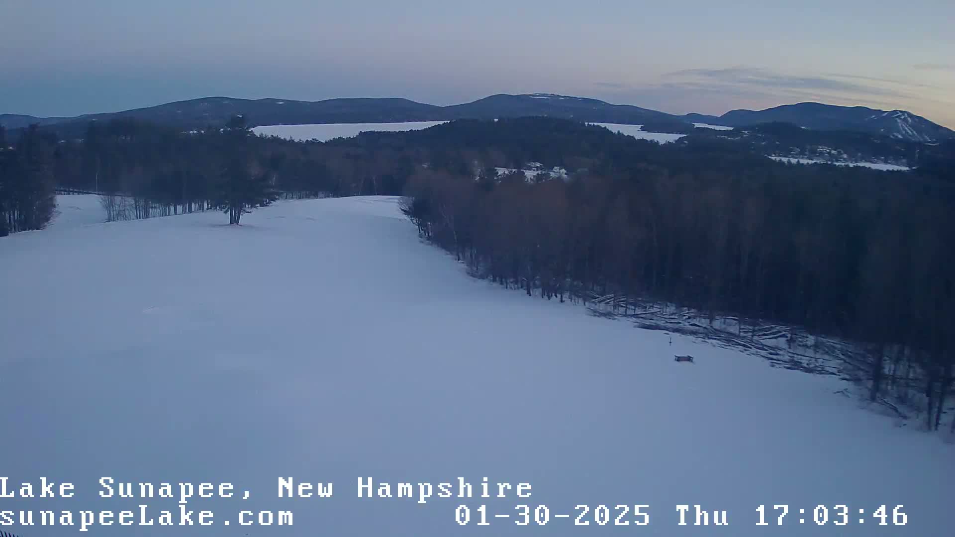 A snow-covered landscape with a lake and mountains in the distance under a twilight sky.